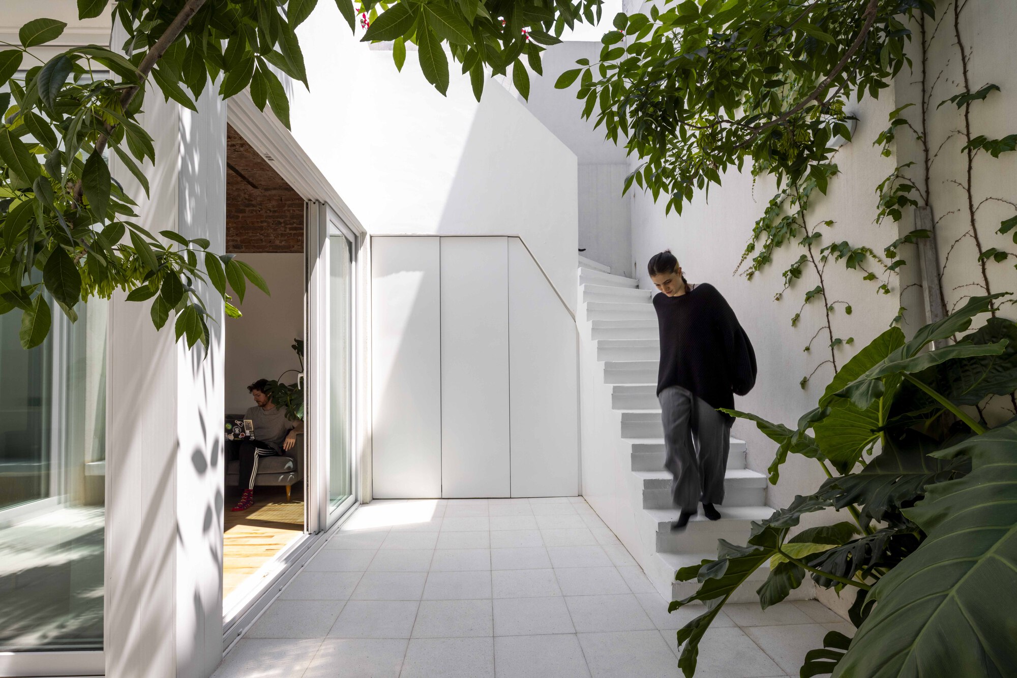 Woman in black walking down the white courtyard stair past large tropical leaves, with a seated man visible through the glass doors