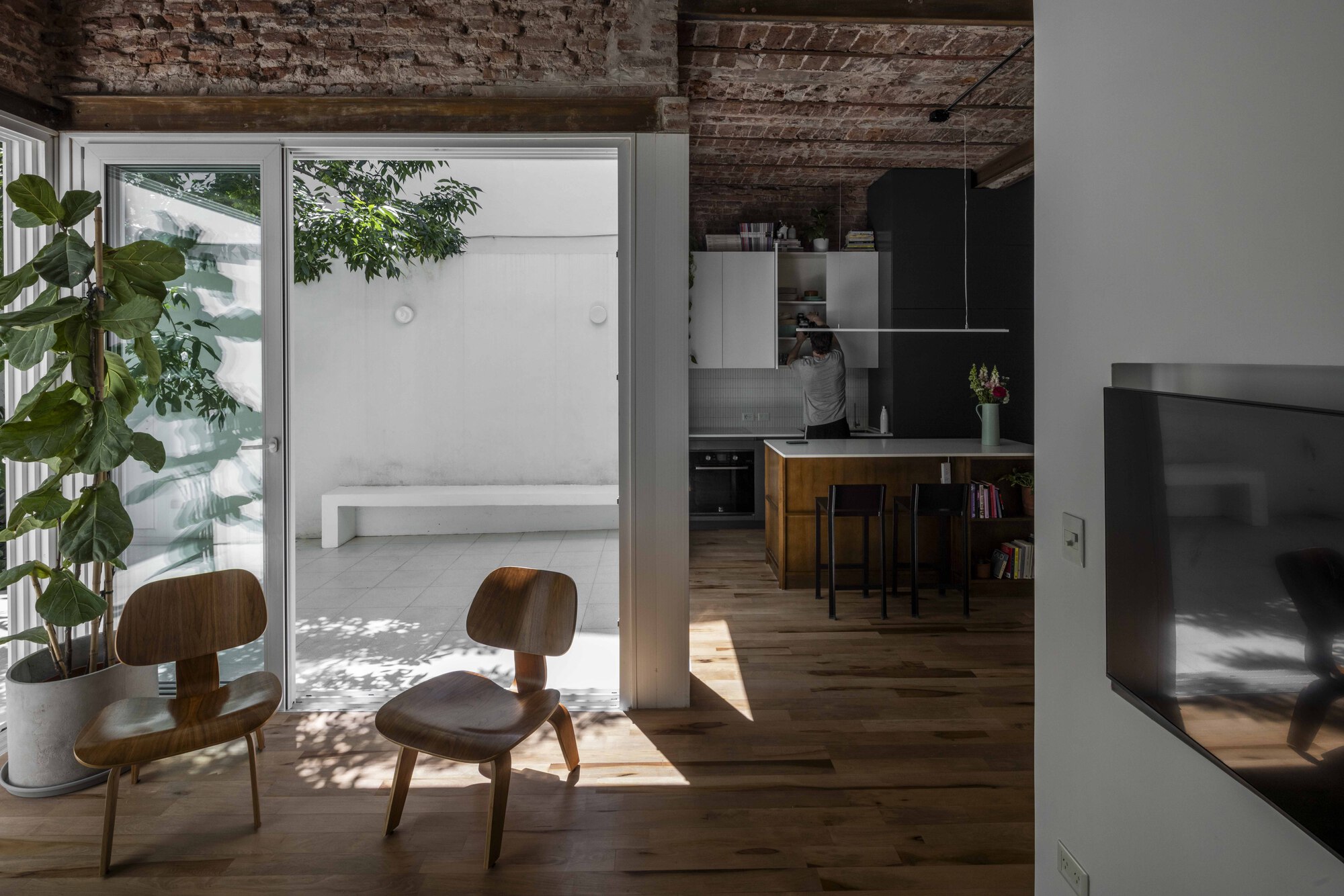 Open sliding glass door linking two plywood lounge chairs to a white patio and kitchen island, with leaf shadows on the floor