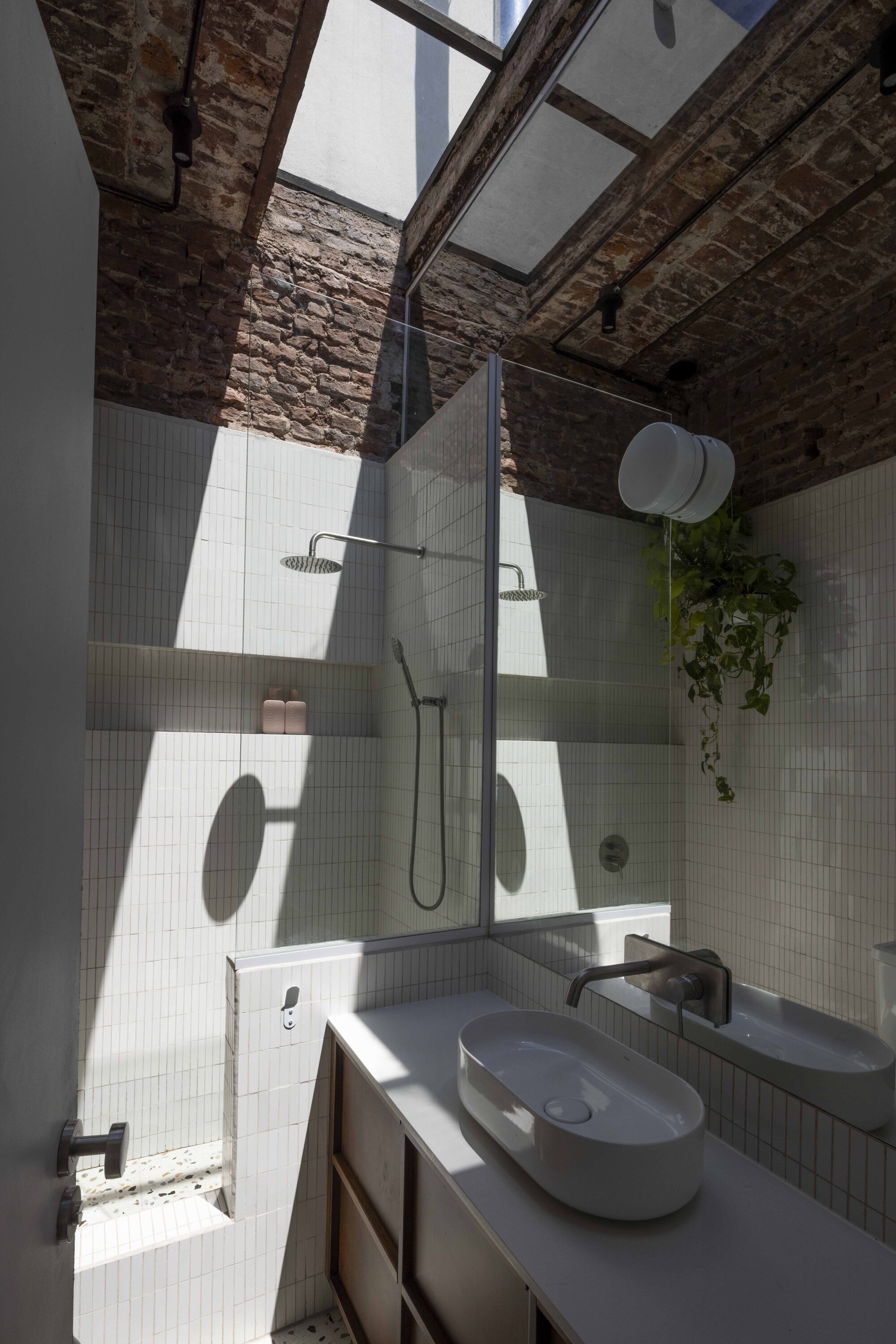 Small bathroom with white tile, a glass shower screen and an oval basin, lit by a skylight cutting through brick