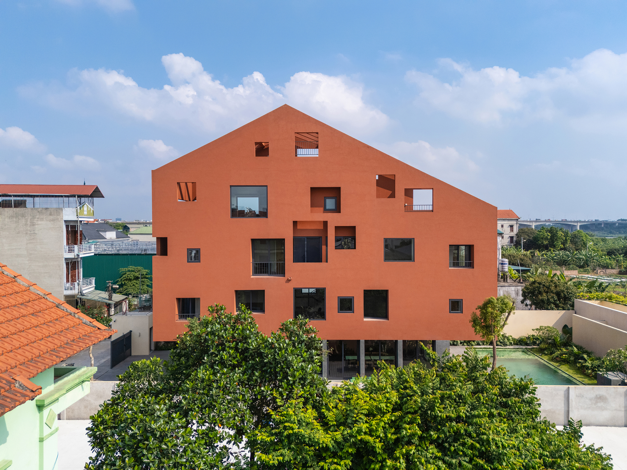 Gabled terracotta-red facade punctured by irregular square windows and recesses, seen over green treetops under a bright blue sky with clouds