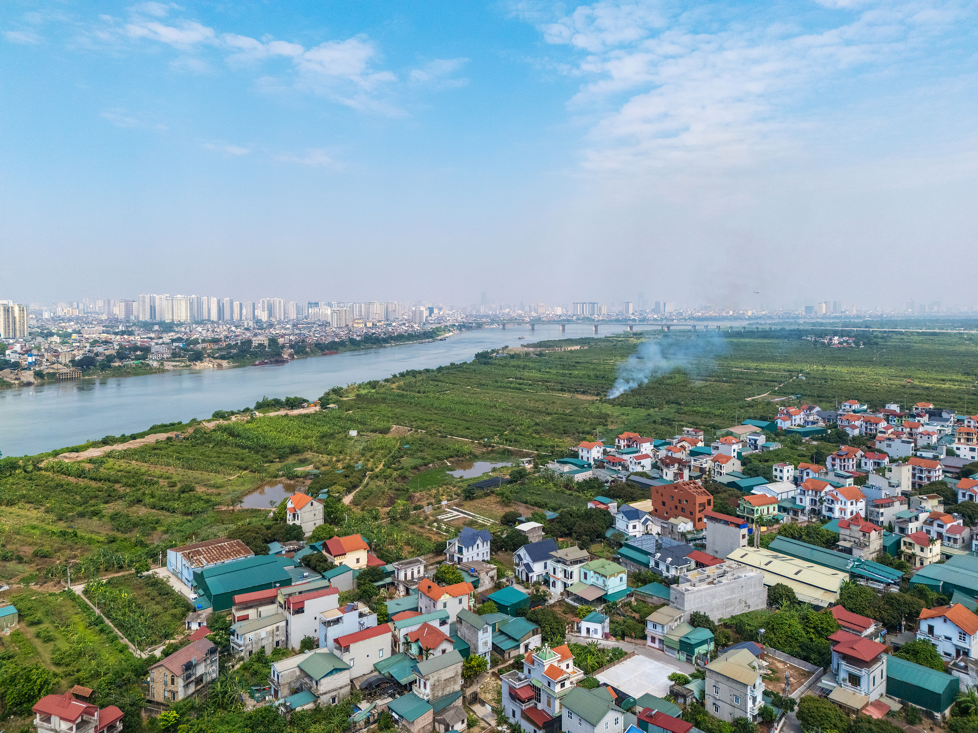 Aerial view of low houses with red and green roofs beside a wide river and farmland, with smoke rising in hazy light