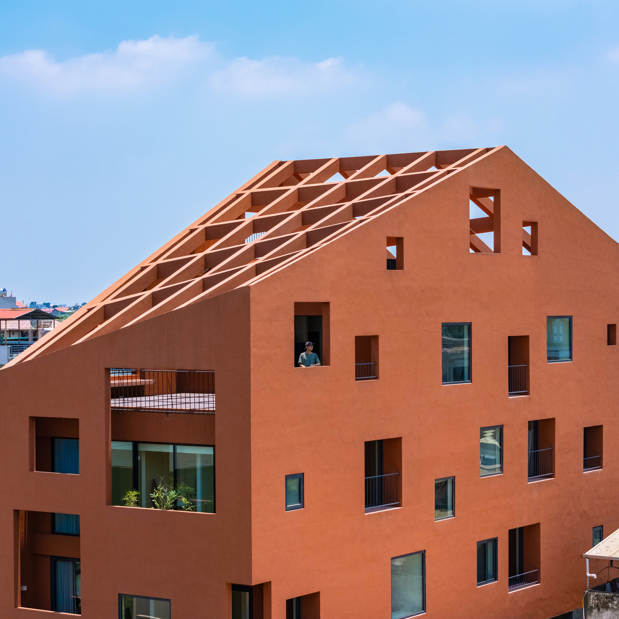 Open timber roof frame above the red gabled facade, with a person leaning out of a recessed window in afternoon sun