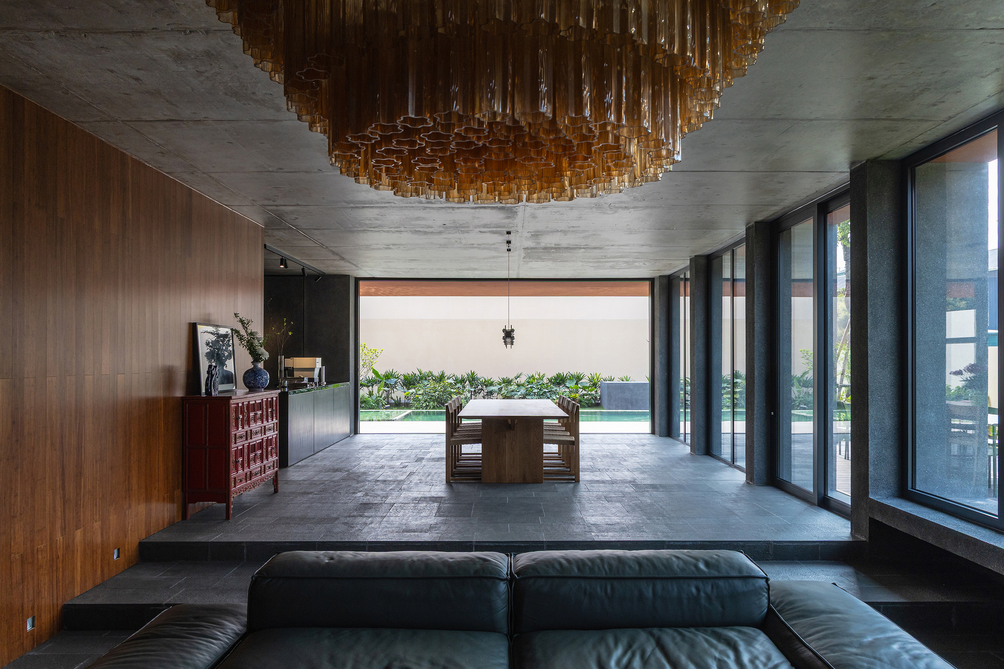 Living and dining space with a stone-tiled platform, amber glass chandelier and timber wall, opening onto a pool and planting