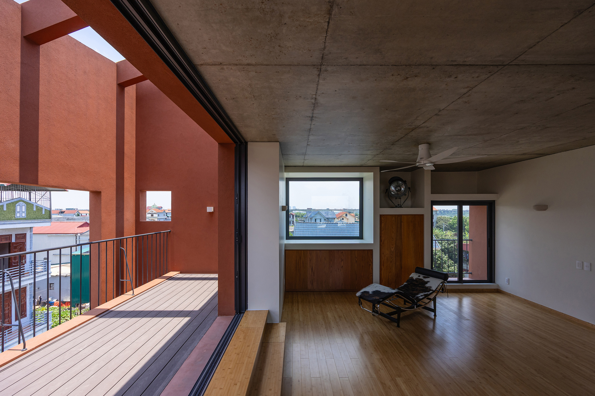 Upper-floor room with bamboo flooring and concrete ceiling, opening through sliding glass onto a decked balcony with a lounge chair inside