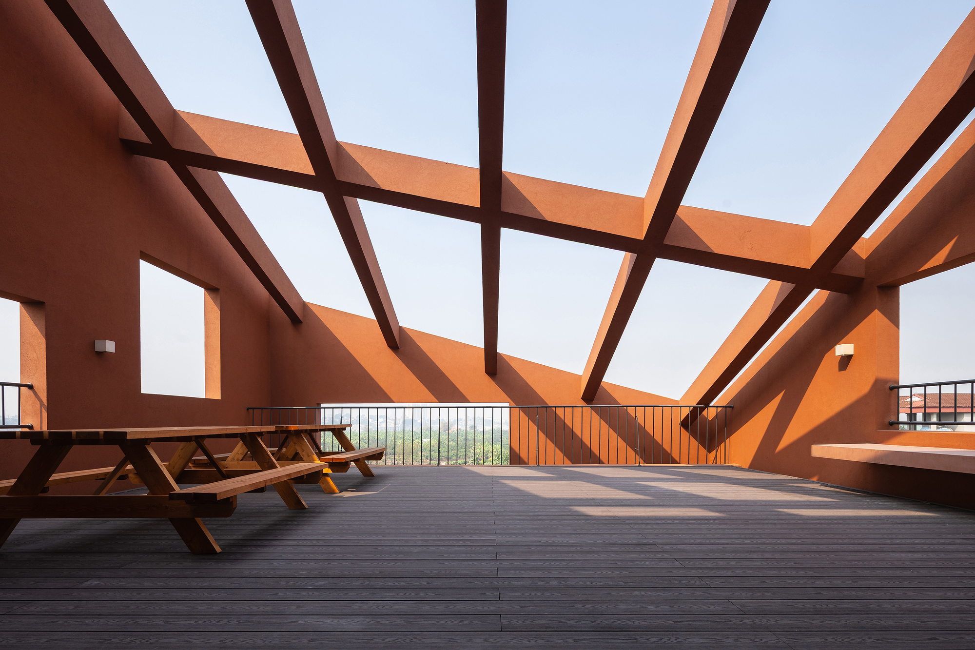 Rooftop terrace under open red beams, with picnic tables on grey decking and hard shadows cast across the walls