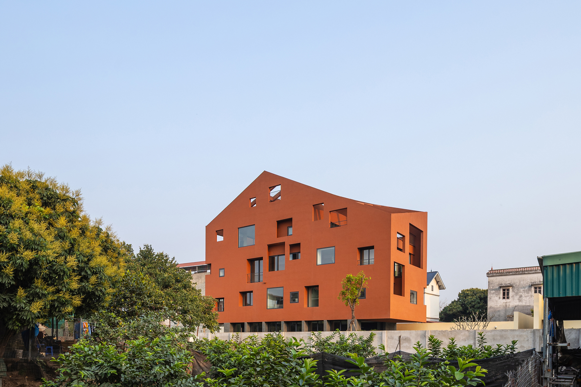Terracotta-orange house with a pitched roofline and scattered window cut-outs, seen over leafy shrubs under a clear afternoon sky