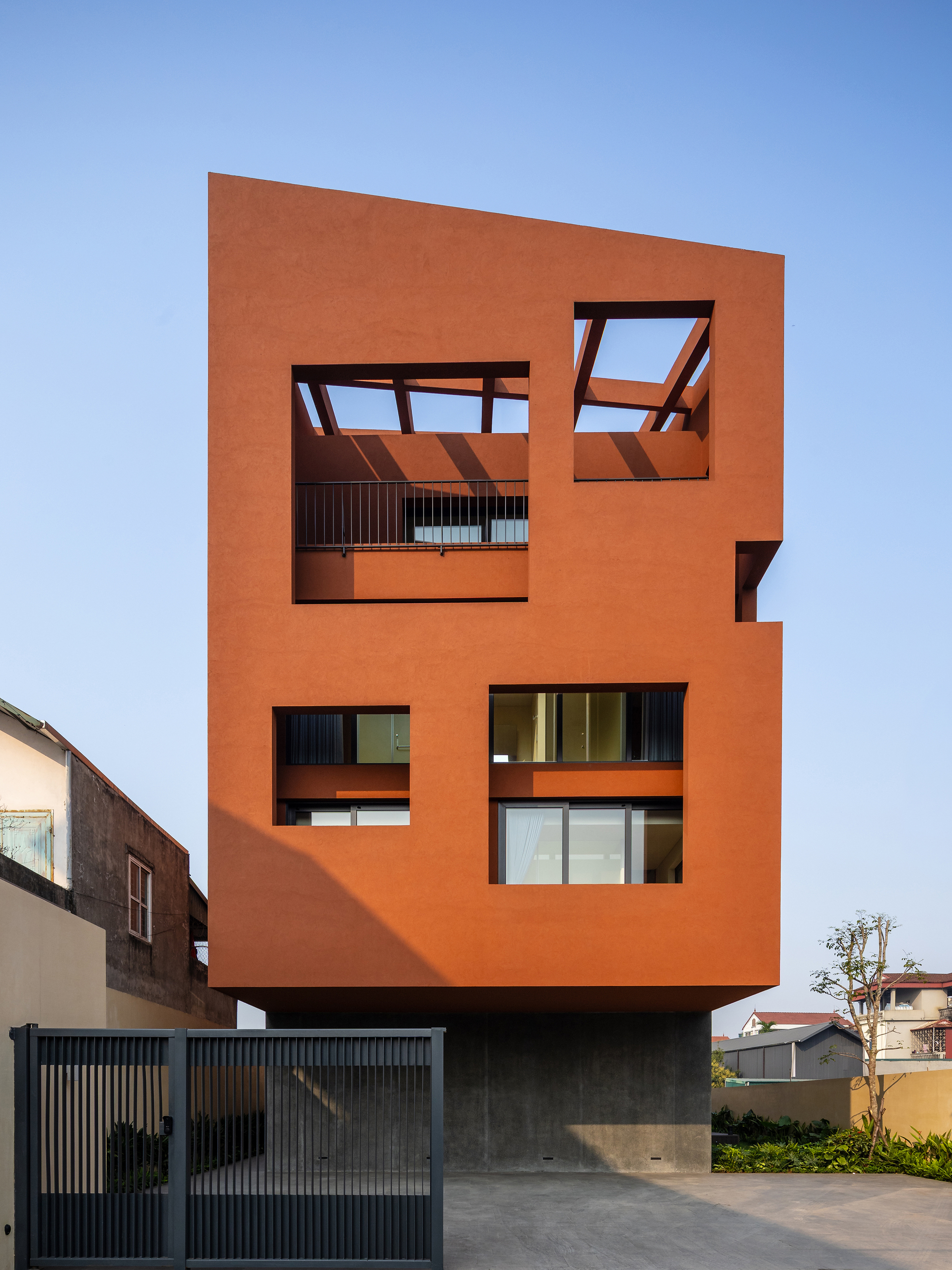 Front facade of the orange volume raised on a concrete base, with open roof frames and a metal gate in blue dusk light