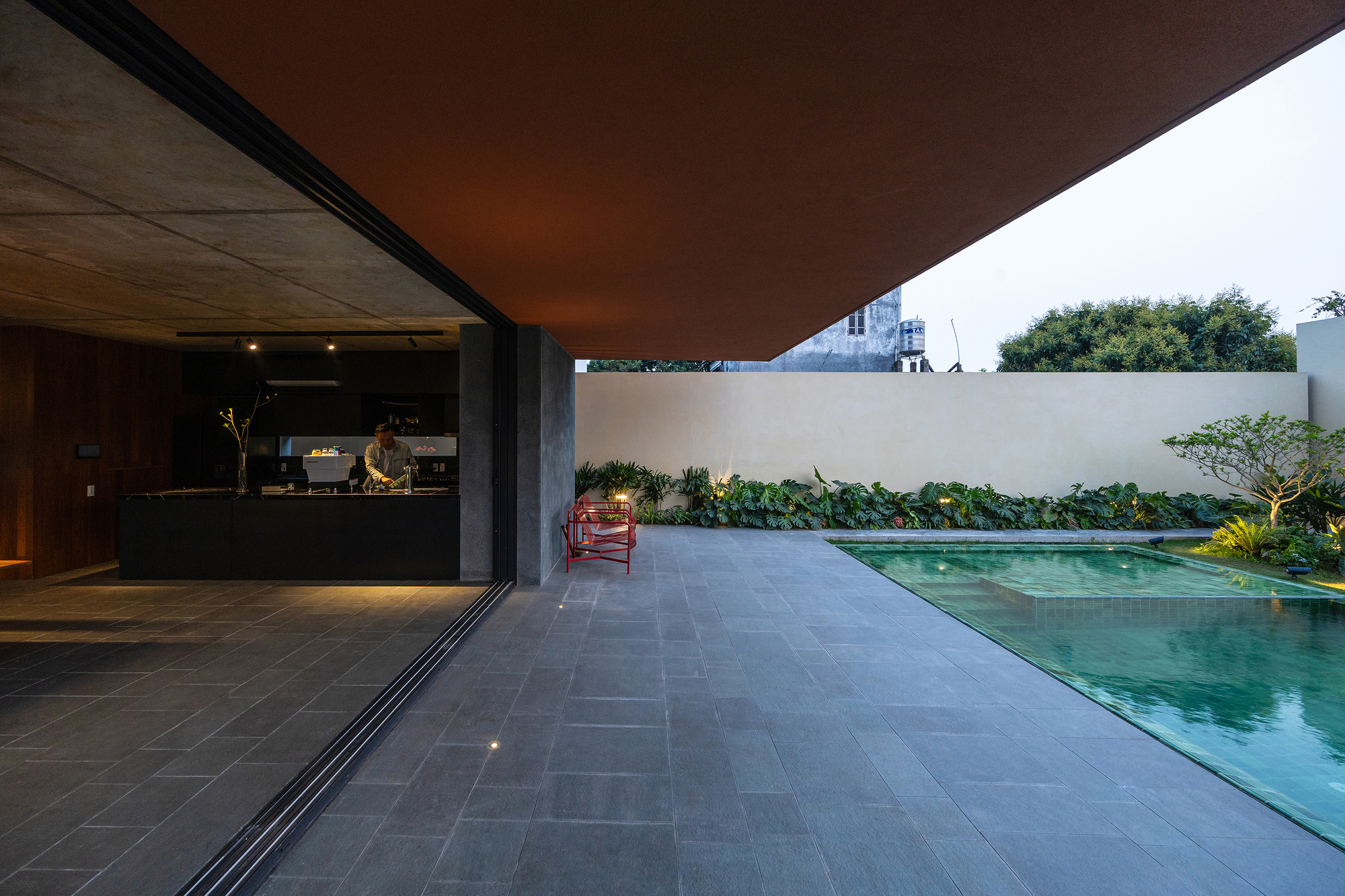 Deep orange soffit overhanging a slate terrace beside the pool, with a person working in the lit kitchen at dusk