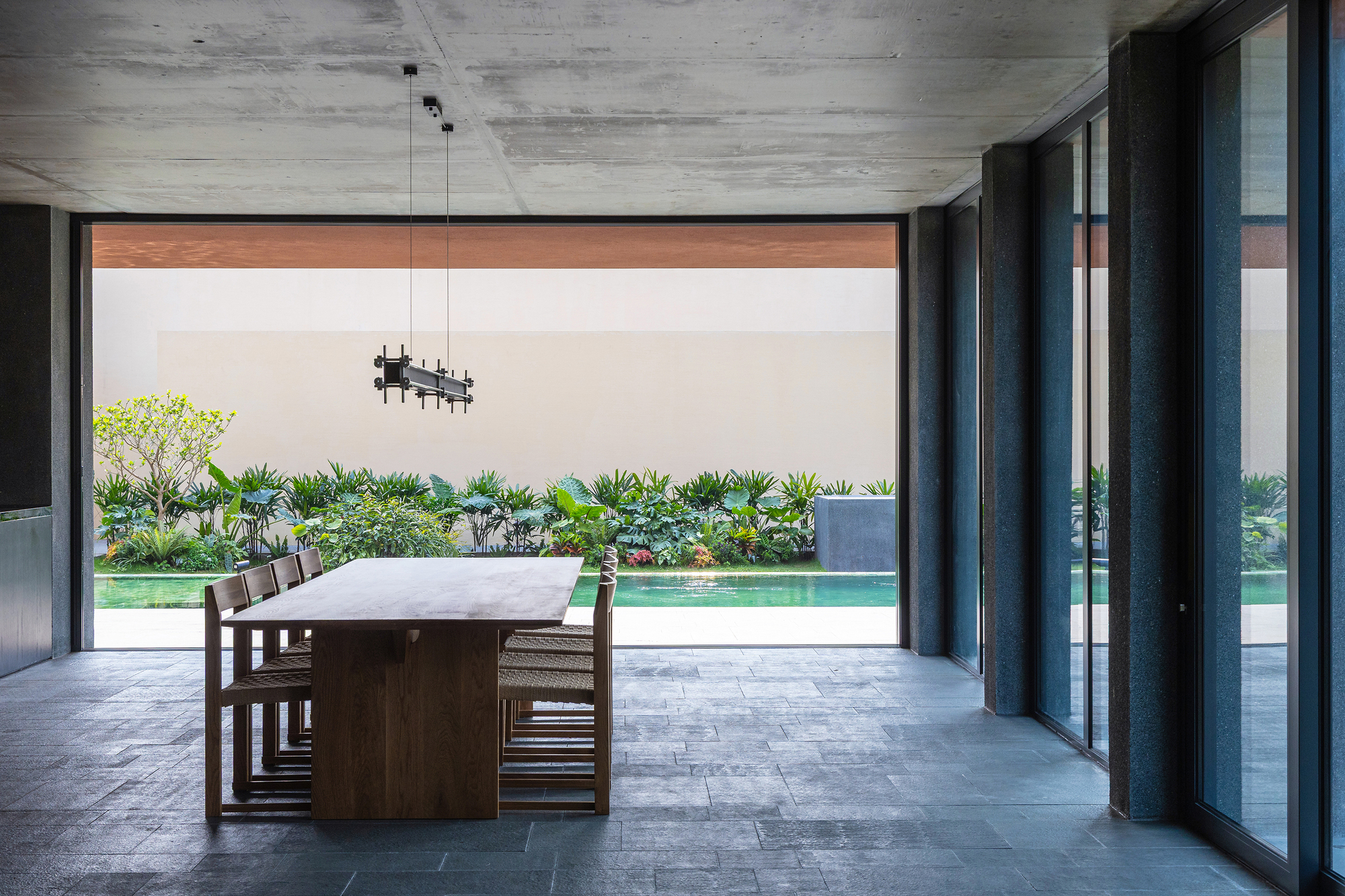 Timber dining table and chairs on a slate floor, opening through glass walls to the pool and planting beyond