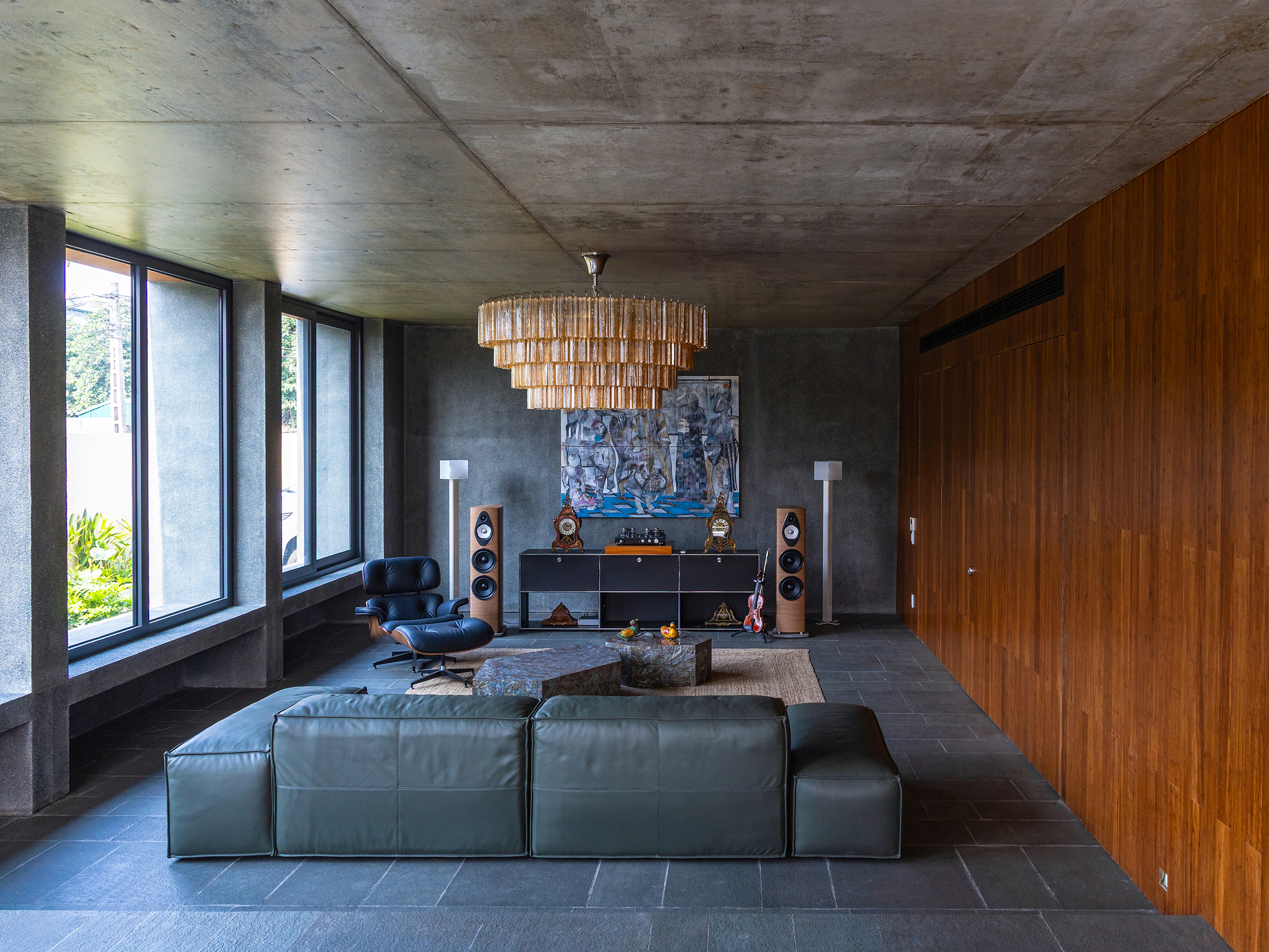 Living room with a green leather sofa, lounge chair and tiered chandelier between a concrete wall and a bamboo-paneled wall