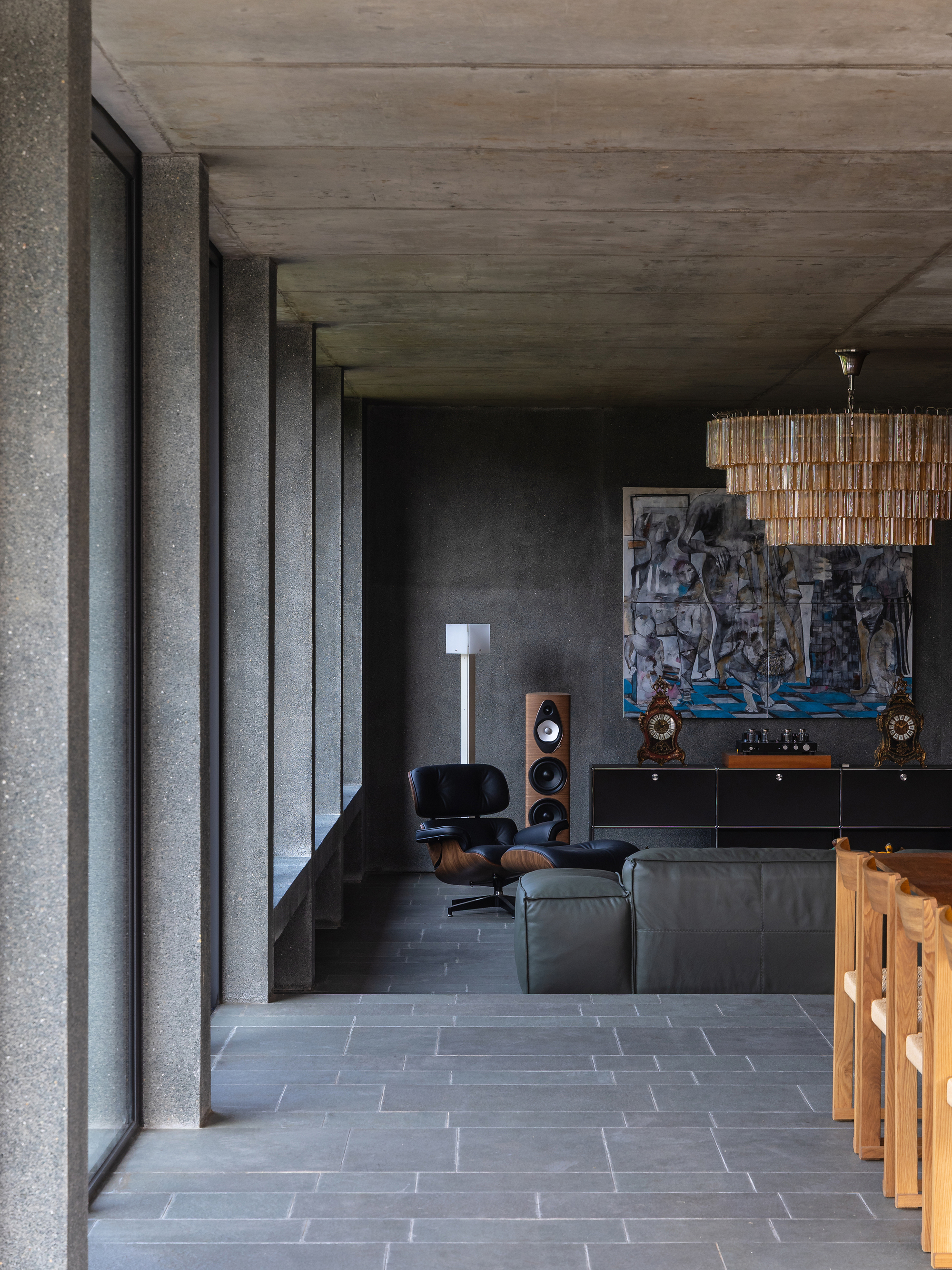 Living area with exposed aggregate concrete columns, slate floor tiles, a black lounge chair and a tiered glass chandelier in low light