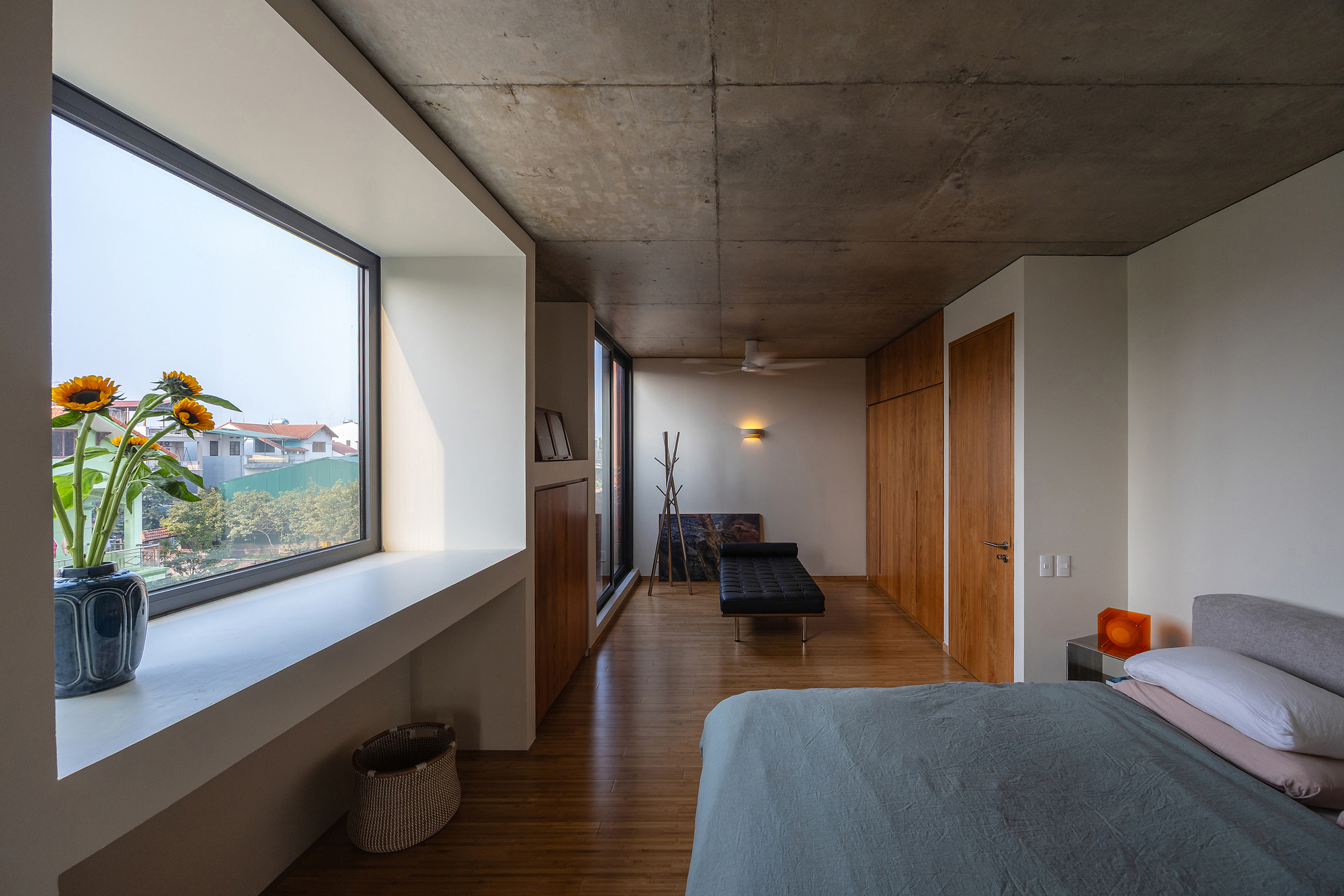 Bedroom with a deep white window recess holding sunflowers, a concrete ceiling and timber wardrobe doors in hazy daylight