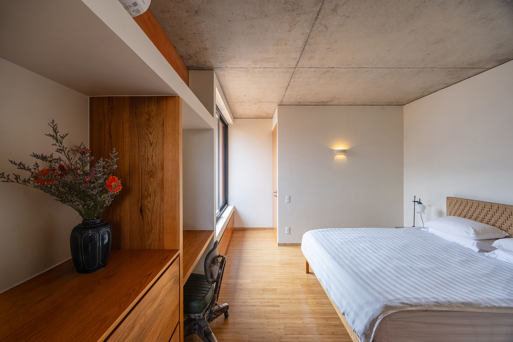 Bedroom with built-in timber shelving, a vase of flowers and a wall sconce glowing against the raw concrete ceiling