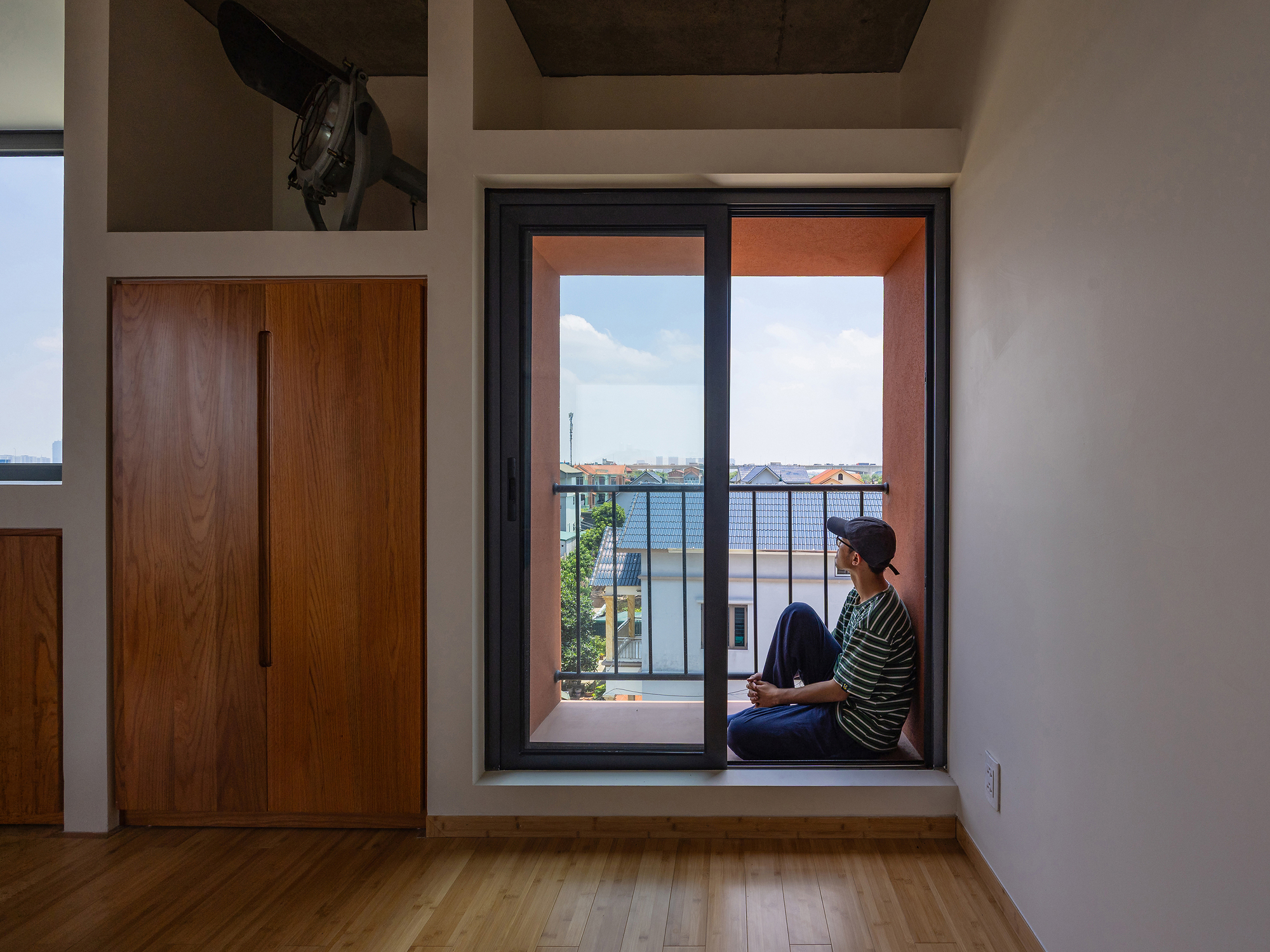Person sitting in a terracotta-walled balcony recess beside a sliding glass door and tall timber cabinet in daylight