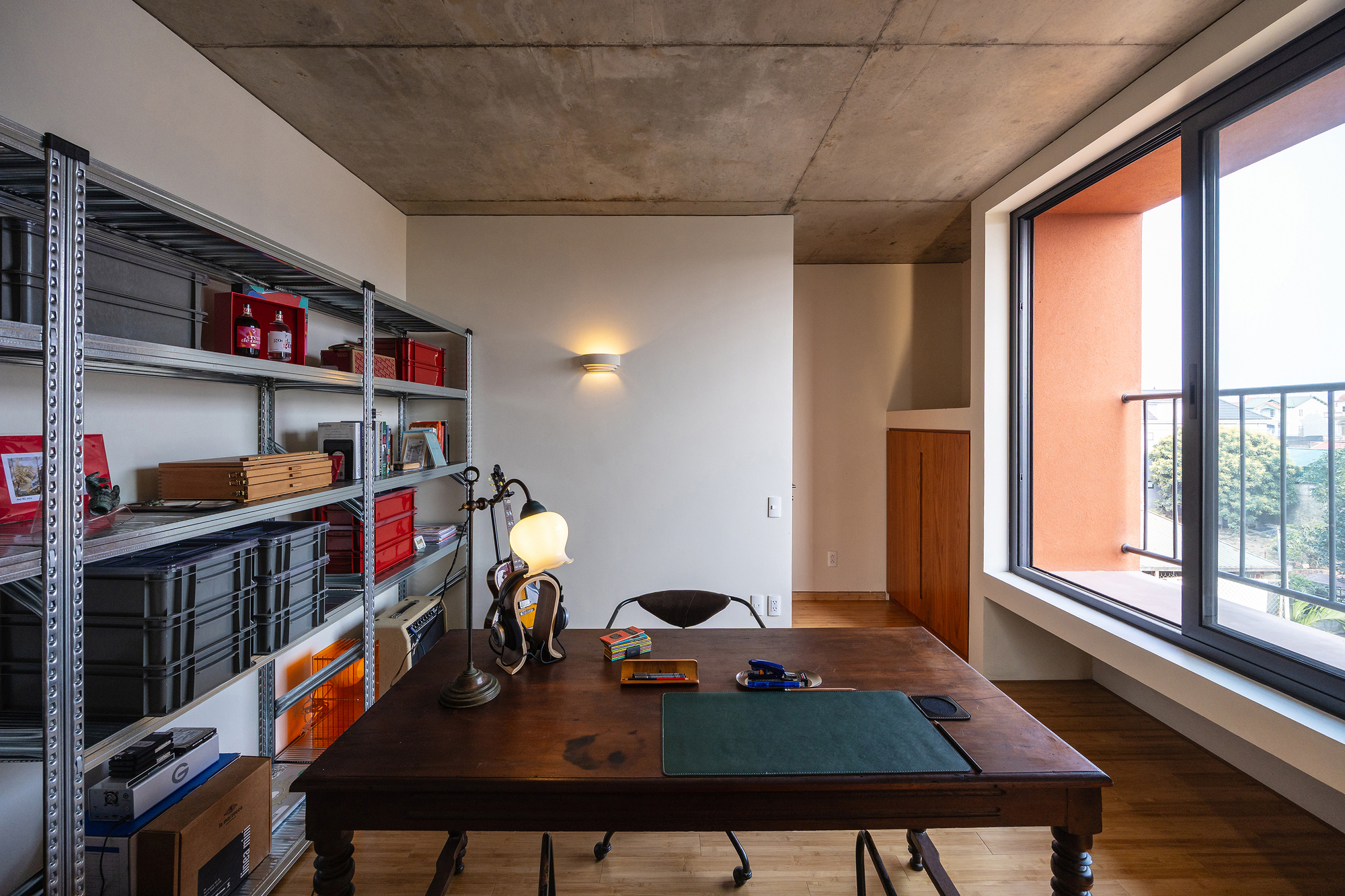 Study with a wooden desk, steel shelving of storage boxes and a bare concrete ceiling beside a window onto a balcony