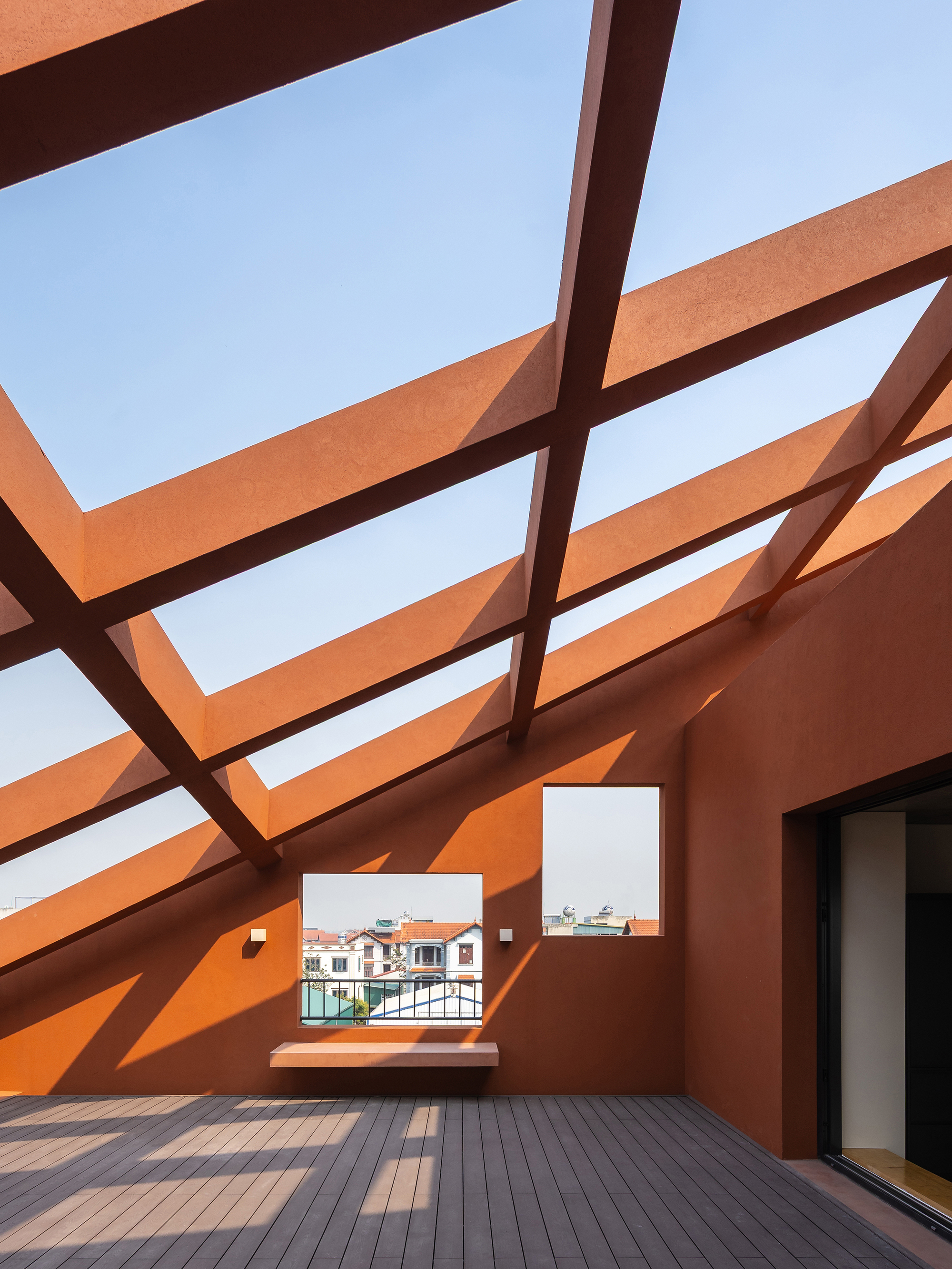 Terracotta-coloured roof terrace with diagonal beams casting striped shadows across a timber deck and a built-in bench under clear sky