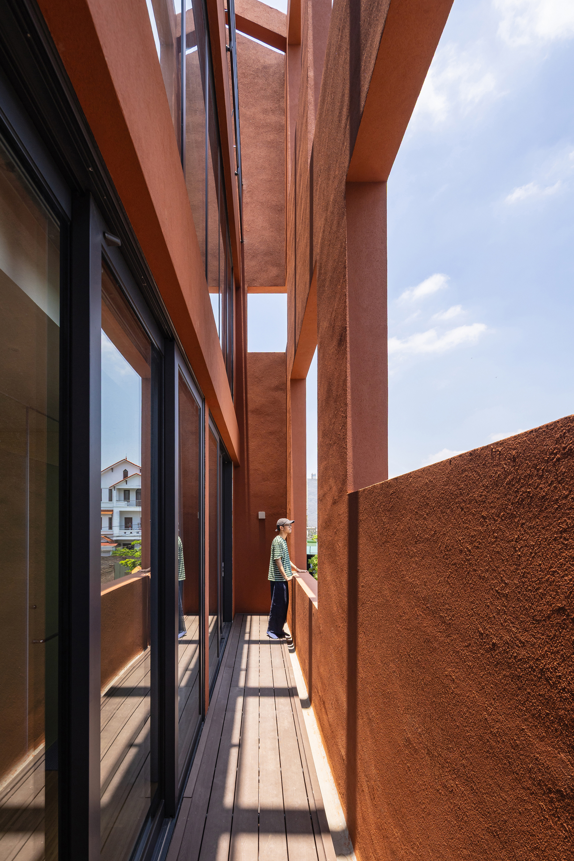 Narrow balcony between glazed sliding doors and a rough terracotta wall, with a person standing at the far end under blue sky