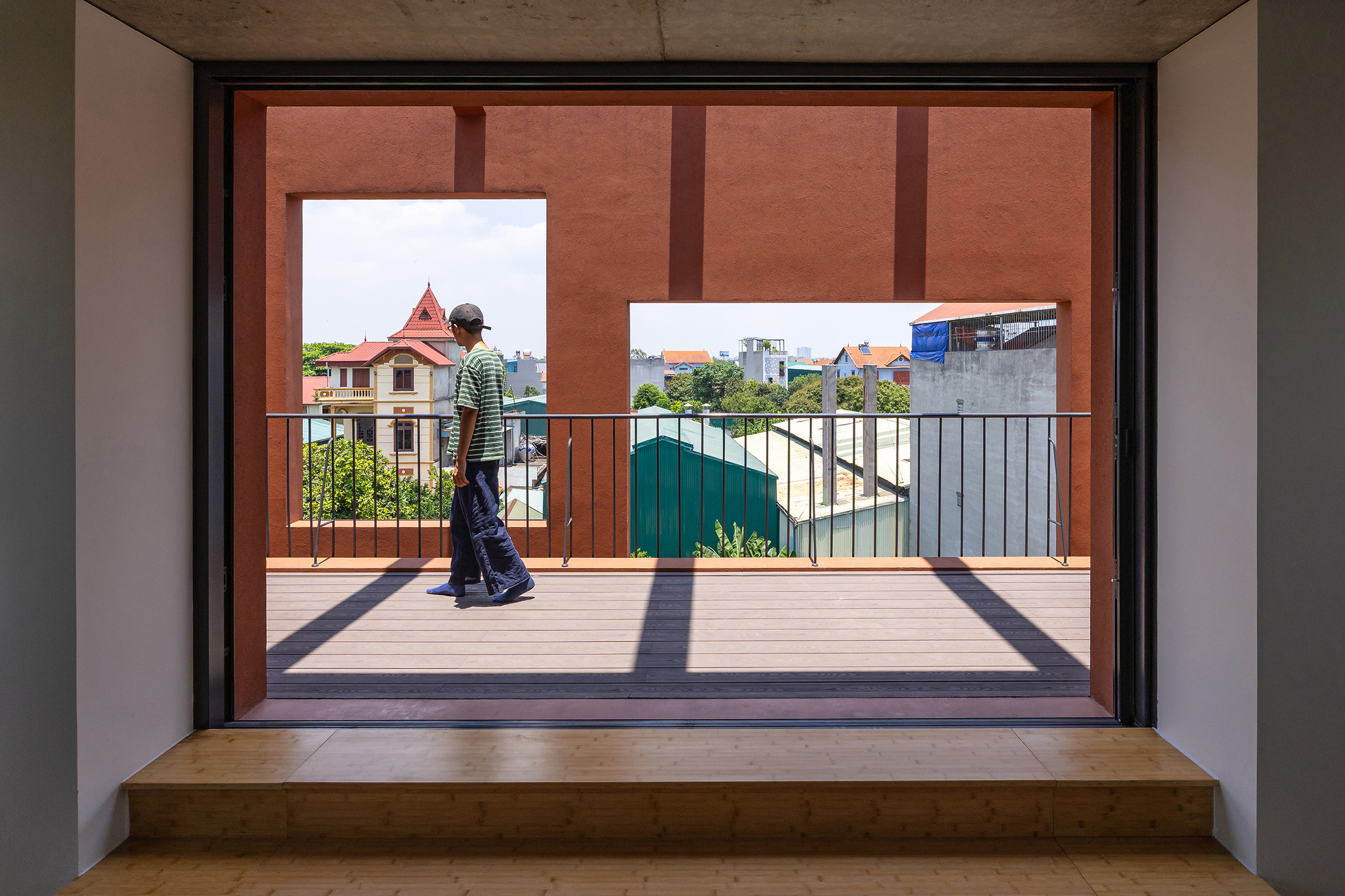 Framed view from a timber-floored interior across a terrace with railing, where a person in a striped shirt walks past terracotta wall openings