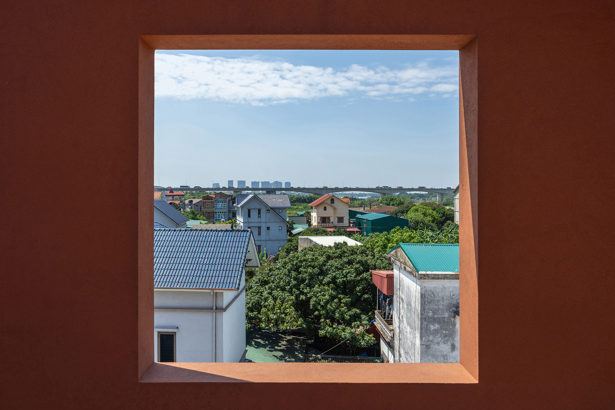 Square window cut into a terracotta-colored wall framing neighboring pitched roofs, a leafy tree and a distant elevated bridge under a blue sky