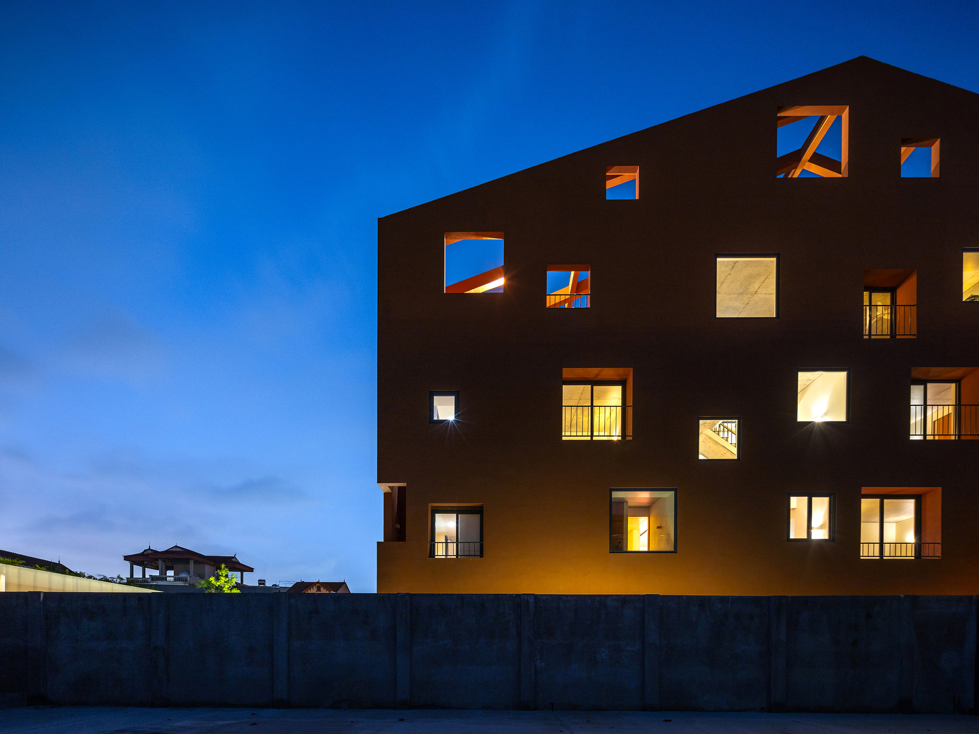 Dusk view of a gabled facade with irregular square openings, some lit from within, rising behind a concrete boundary wall