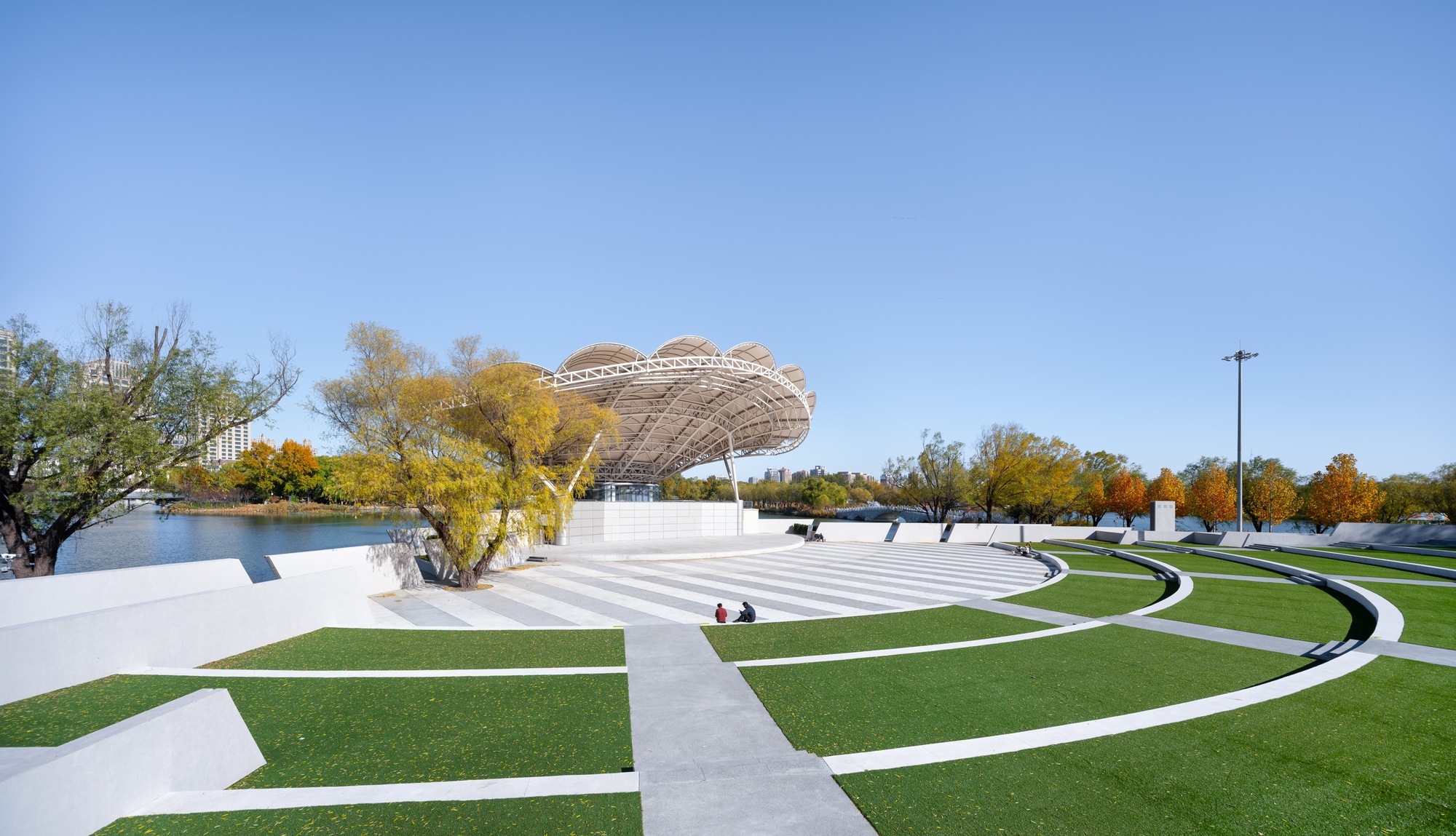 Terraced grass amphitheater with curved white edges facing a stage under a scalloped steel canopy, beside a lake in autumn color