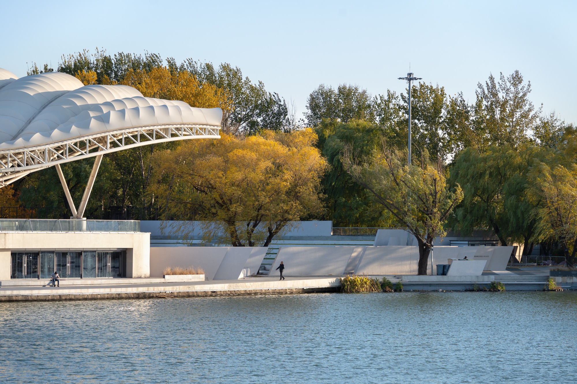 Lakeside stage canopy with quilted white membrane on a steel truss, a lone walker on the waterfront in low golden light