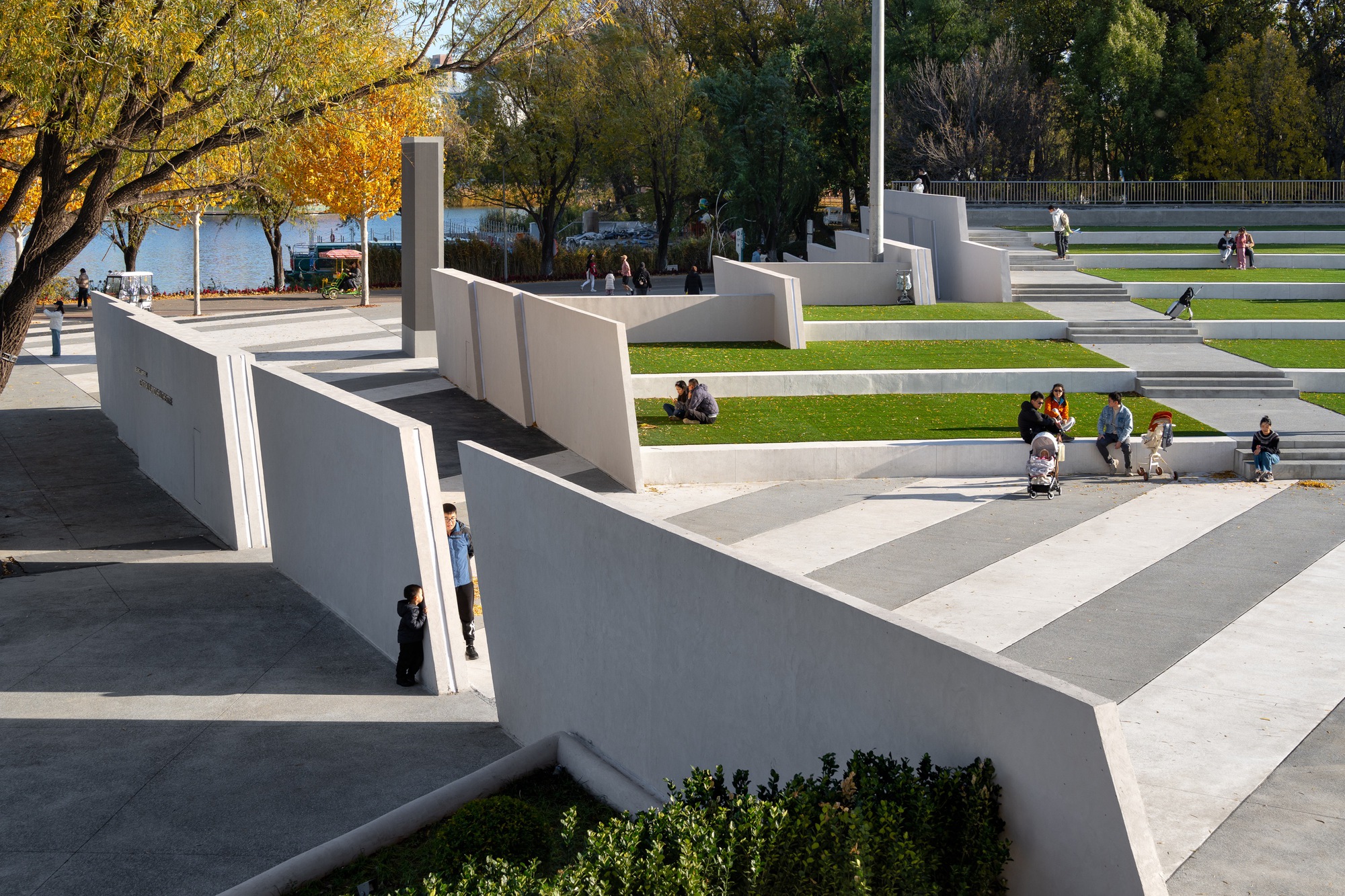 Raised view of angled white walls and striped paving beside grass terraces, with families sitting on the steps in afternoon sun