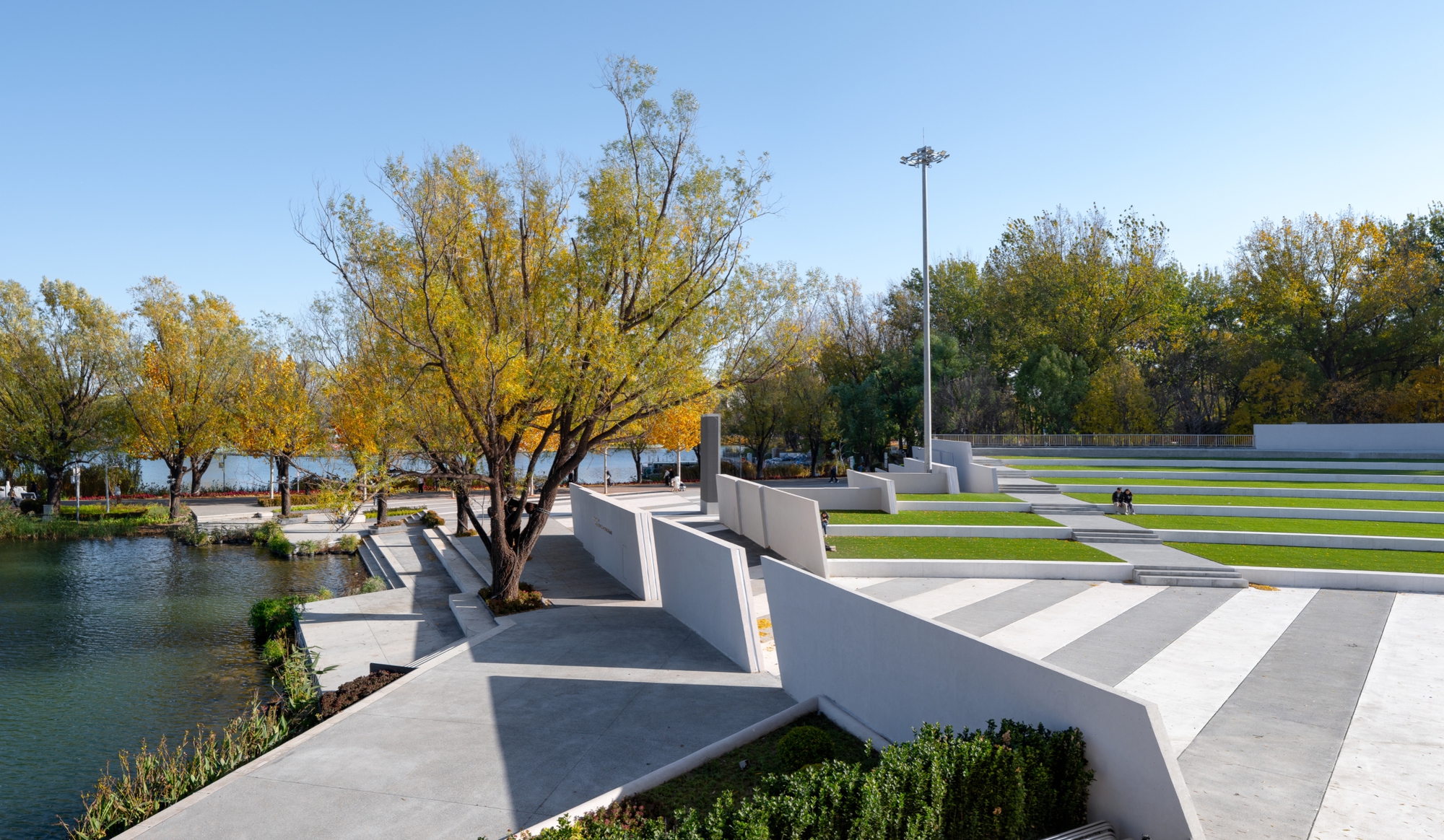 Striped paving and slanted white walls stepping down toward the pond, with autumn willows and a tall light pole overhead