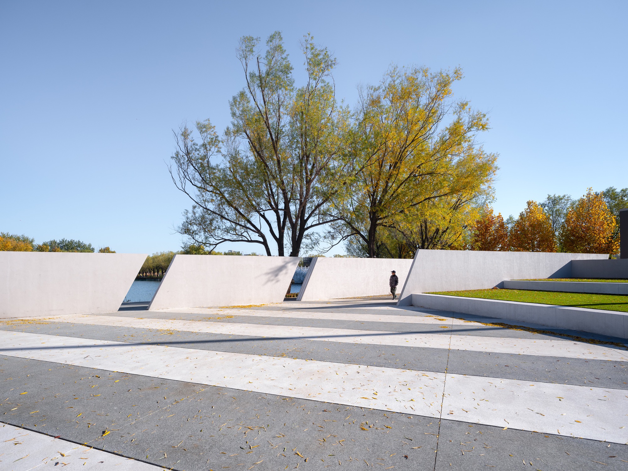 Row of tilted white concrete walls over striped paving with a pedestrian passing, willows and lake glimpsed through the openings under clear sky