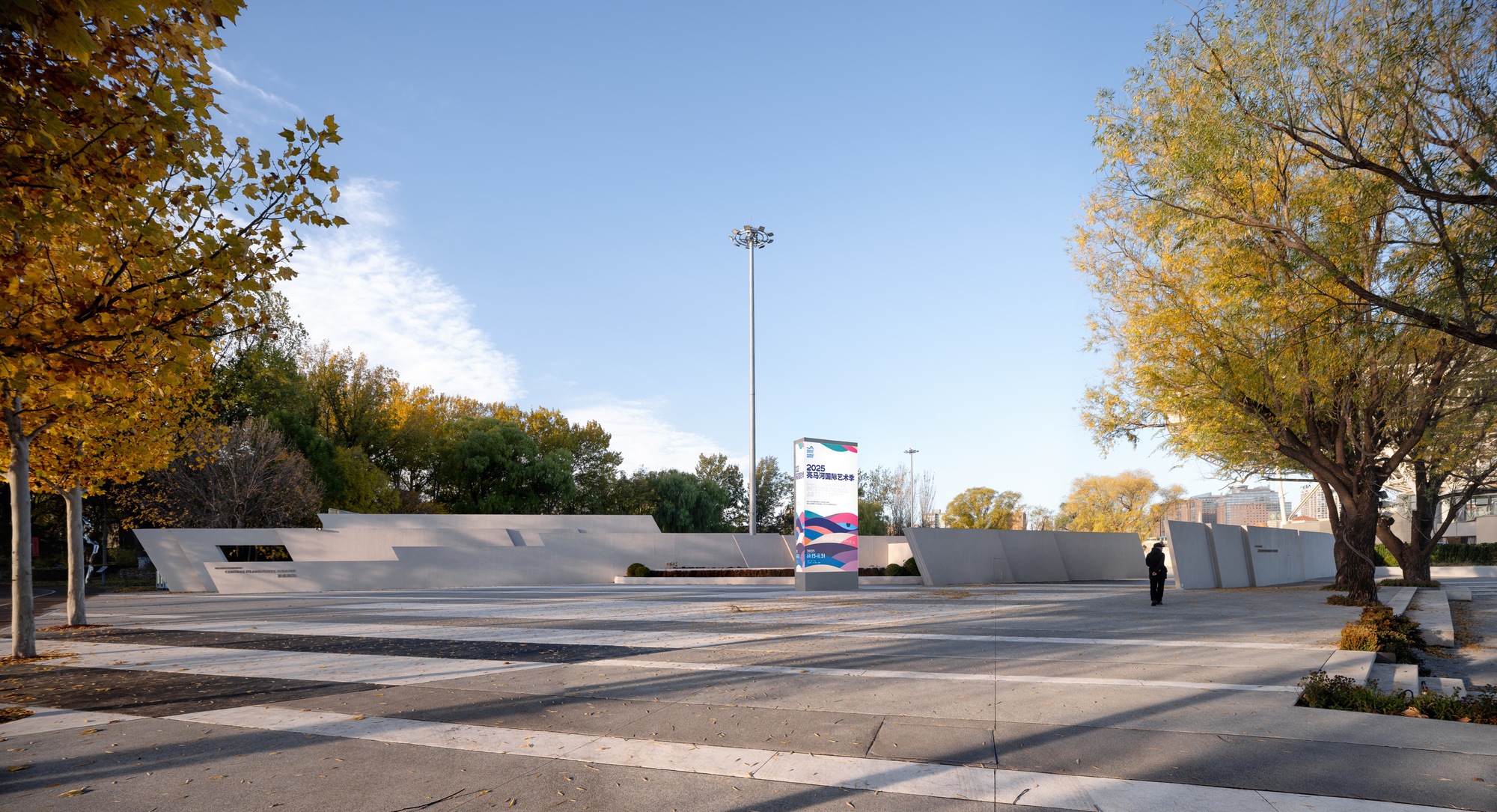 Angled concrete wall segments around a paved plaza with a tall light mast and a banner sign, under yellow autumn trees