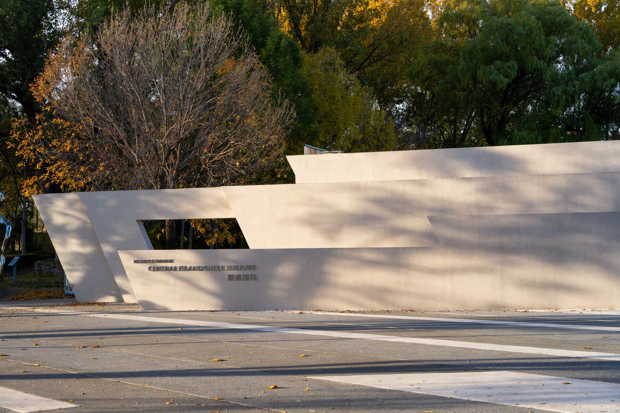 Close view of the stepped, slanted white wall with a rectangular opening and lettering, dappled by tree shadows in warm light