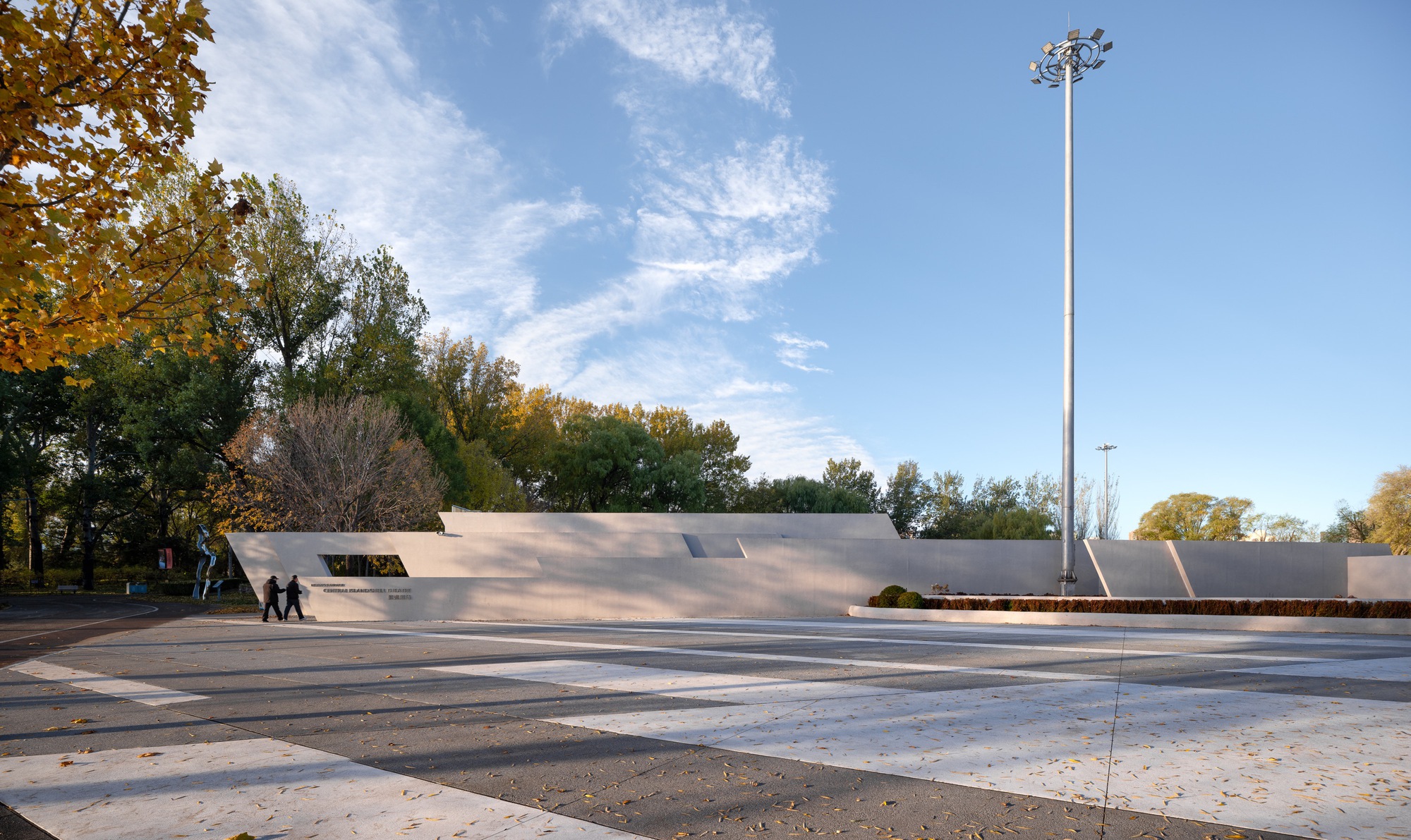 Wide view of the low angular wall along a striped paving plaza, with a tall light pole and wispy clouds
