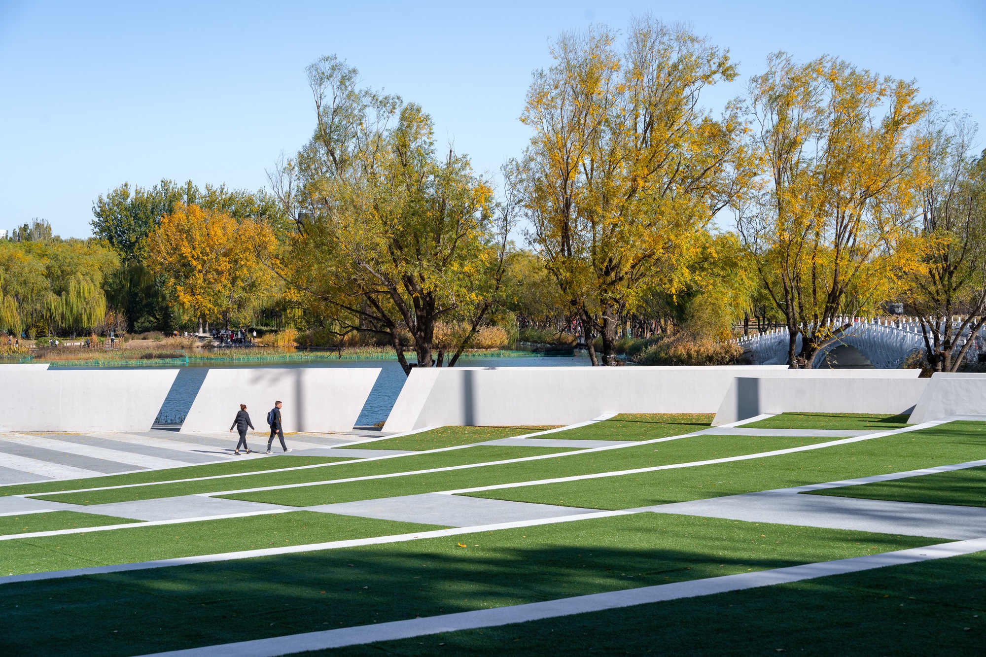 Terraced lawn with white paving bands and angled concrete walls above a lake, with two walkers and yellowing trees