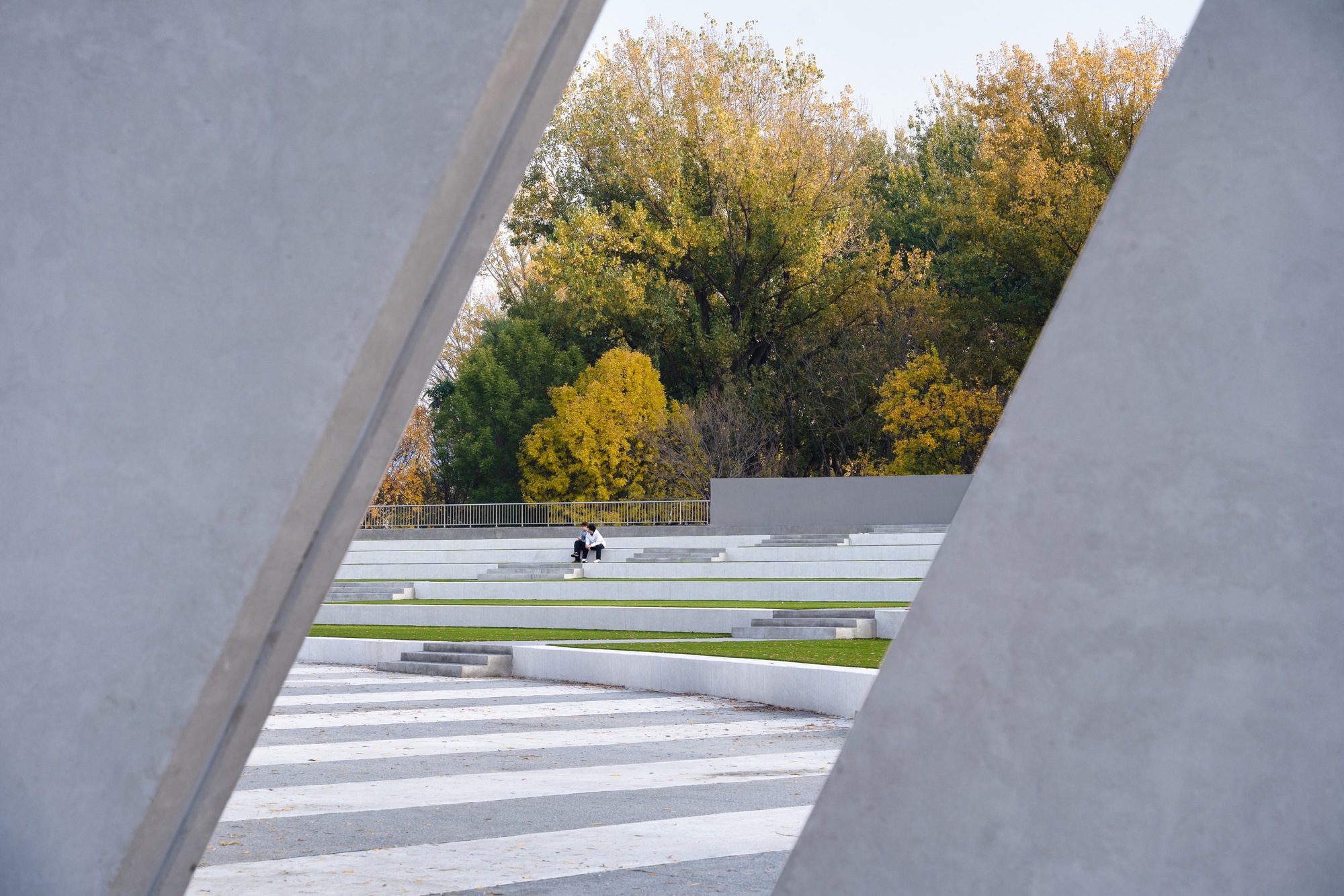 View between two slanted concrete slabs onto striped paving, grass-edged stepped seating and a seated person under autumn foliage
