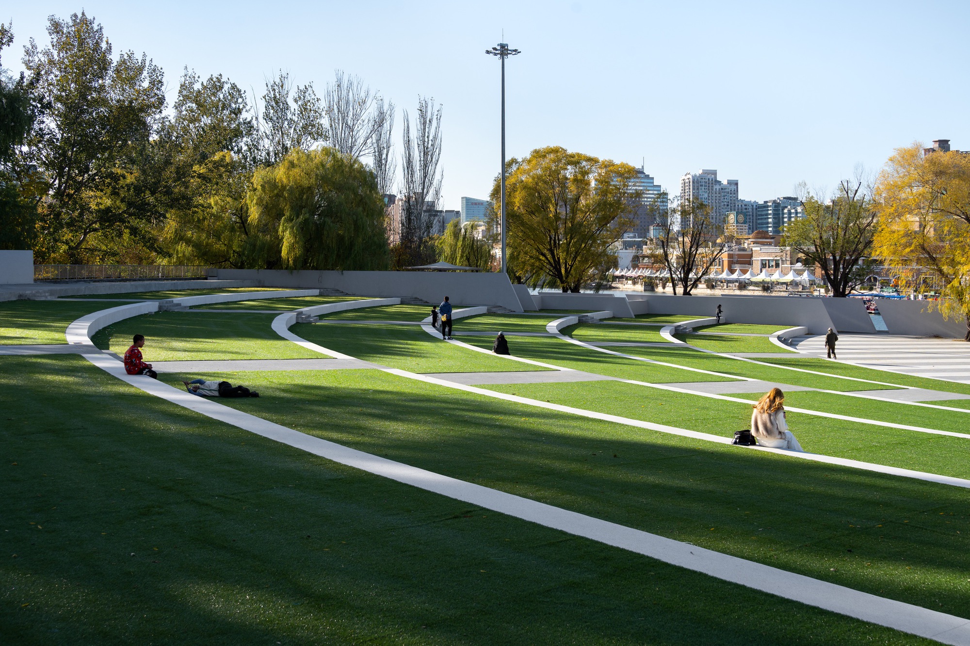 Curving white bands cutting across a grass amphitheater slope, with people sitting on the lawn beneath yellowing trees