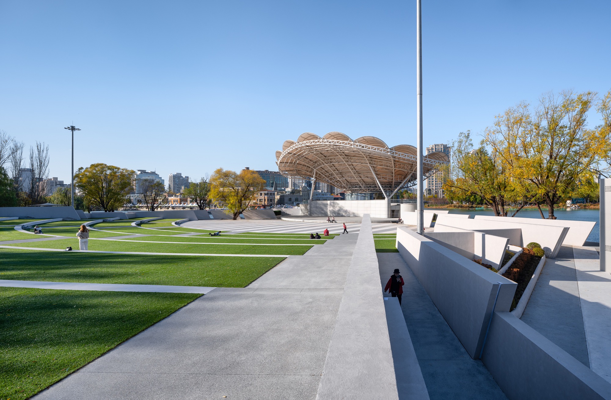 Concrete ramp and angled walls beside the lawn terraces, with the lattice canopy of the stage rising in the distance under clear sky