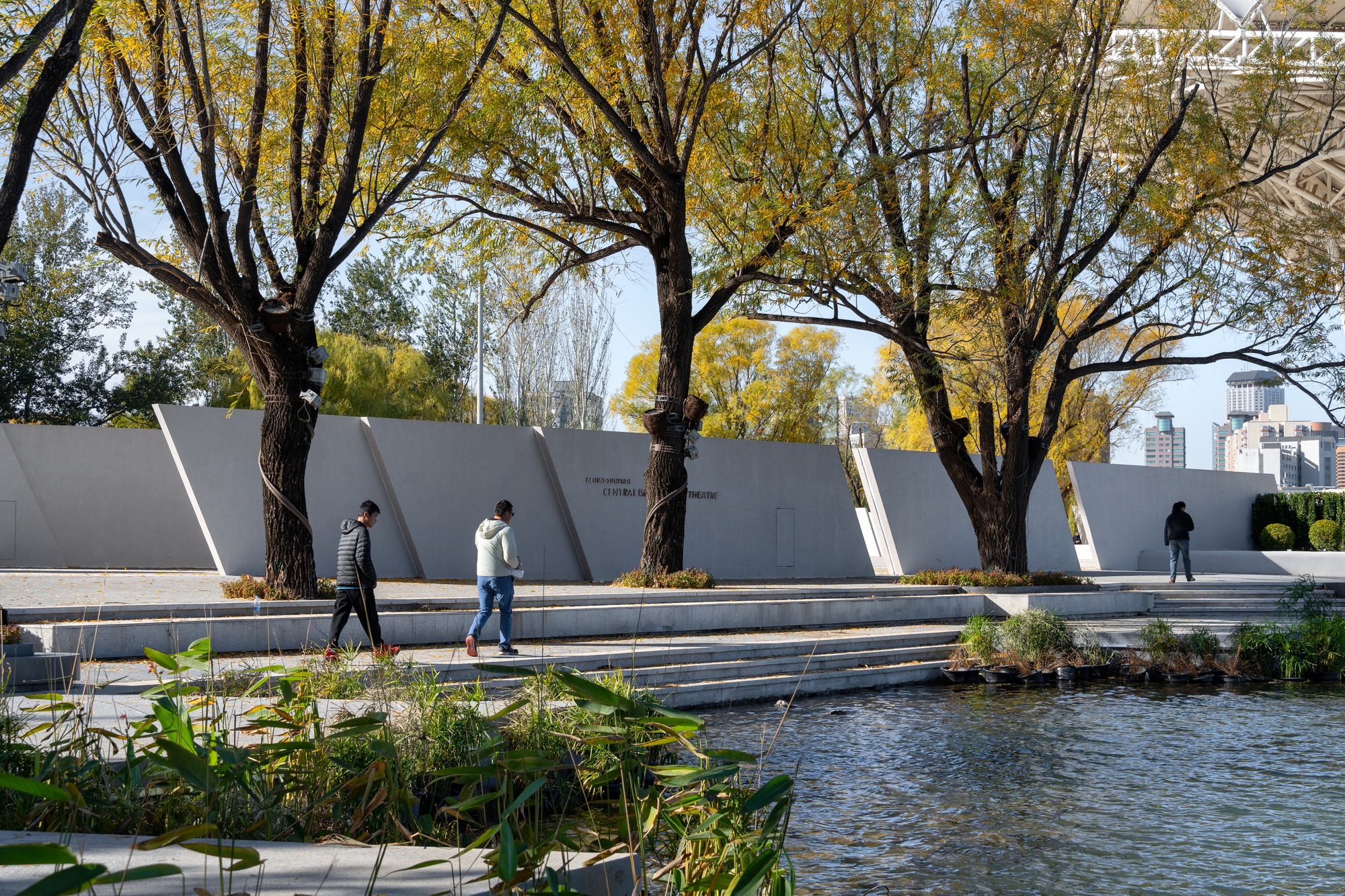 Row of angled concrete wall panels behind stepped edges and a planted pond, with two walkers passing under autumn willows
