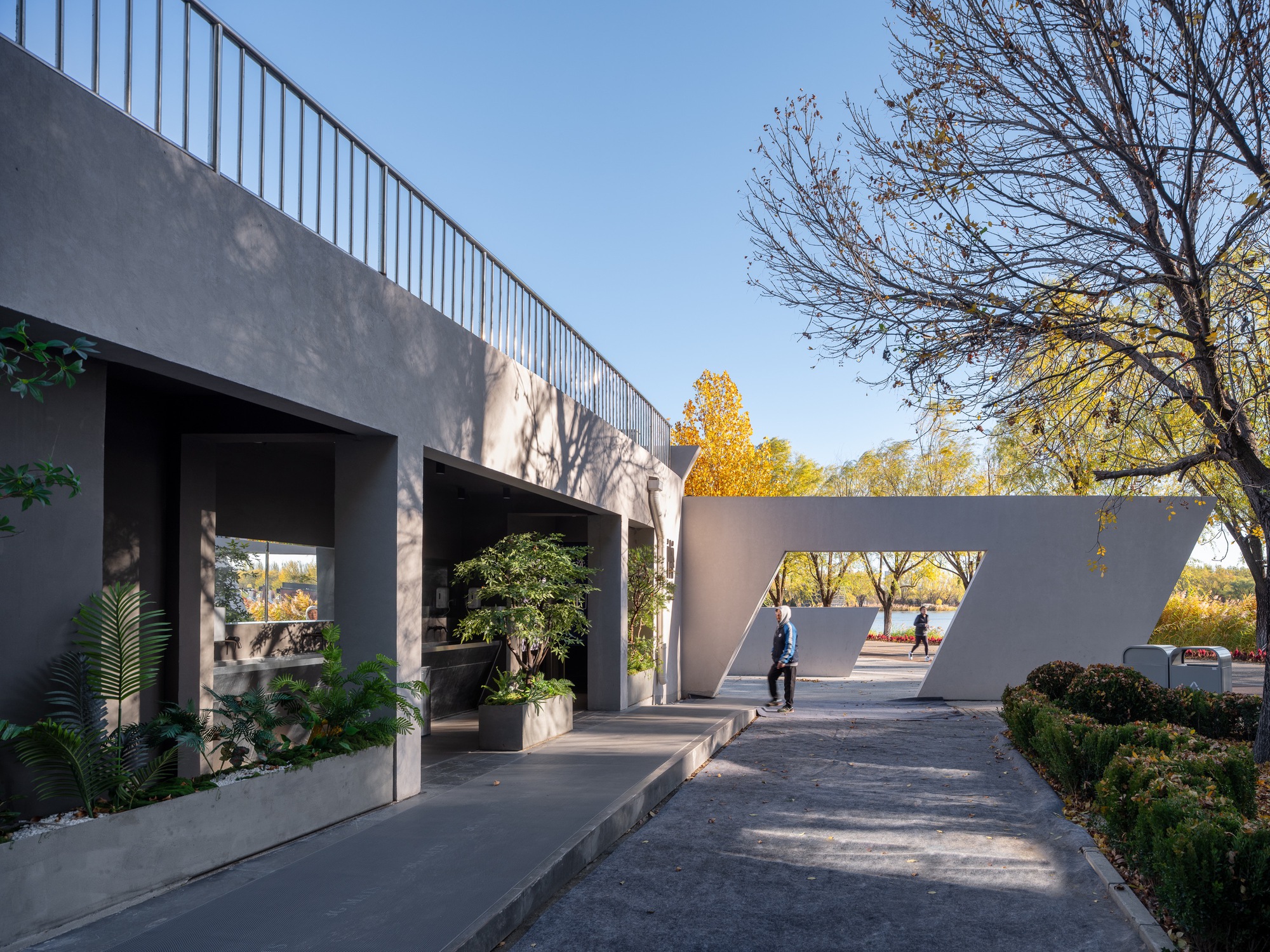 Grey covered walkway with square columns and potted plants beside an open angular frame, a passerby walking in afternoon light