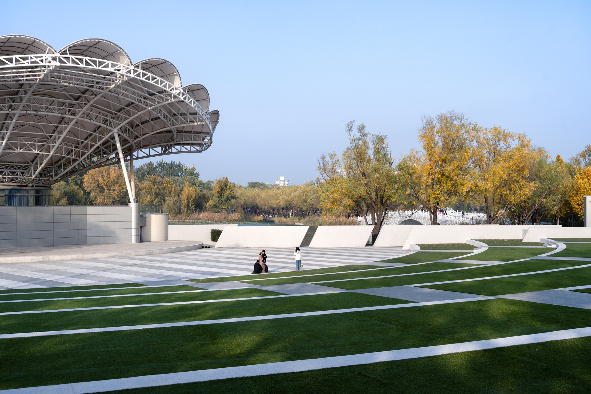 Scalloped steel-truss canopy beside a striped lawn amphitheatre with seated visitors, framed by yellow autumn trees