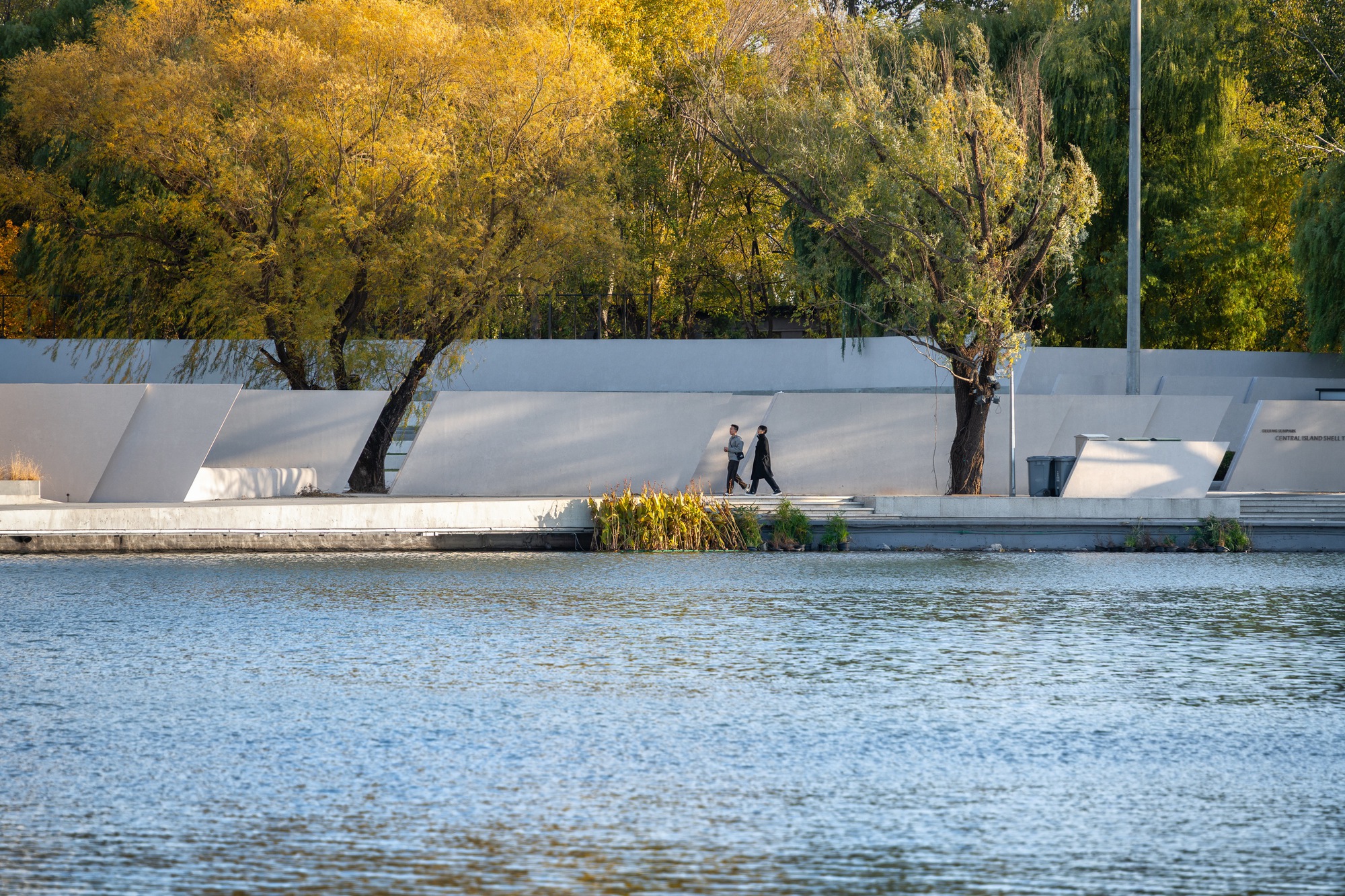 Angled white concrete walls along a lakeside edge, two people walking past willows turning yellow in low autumn light