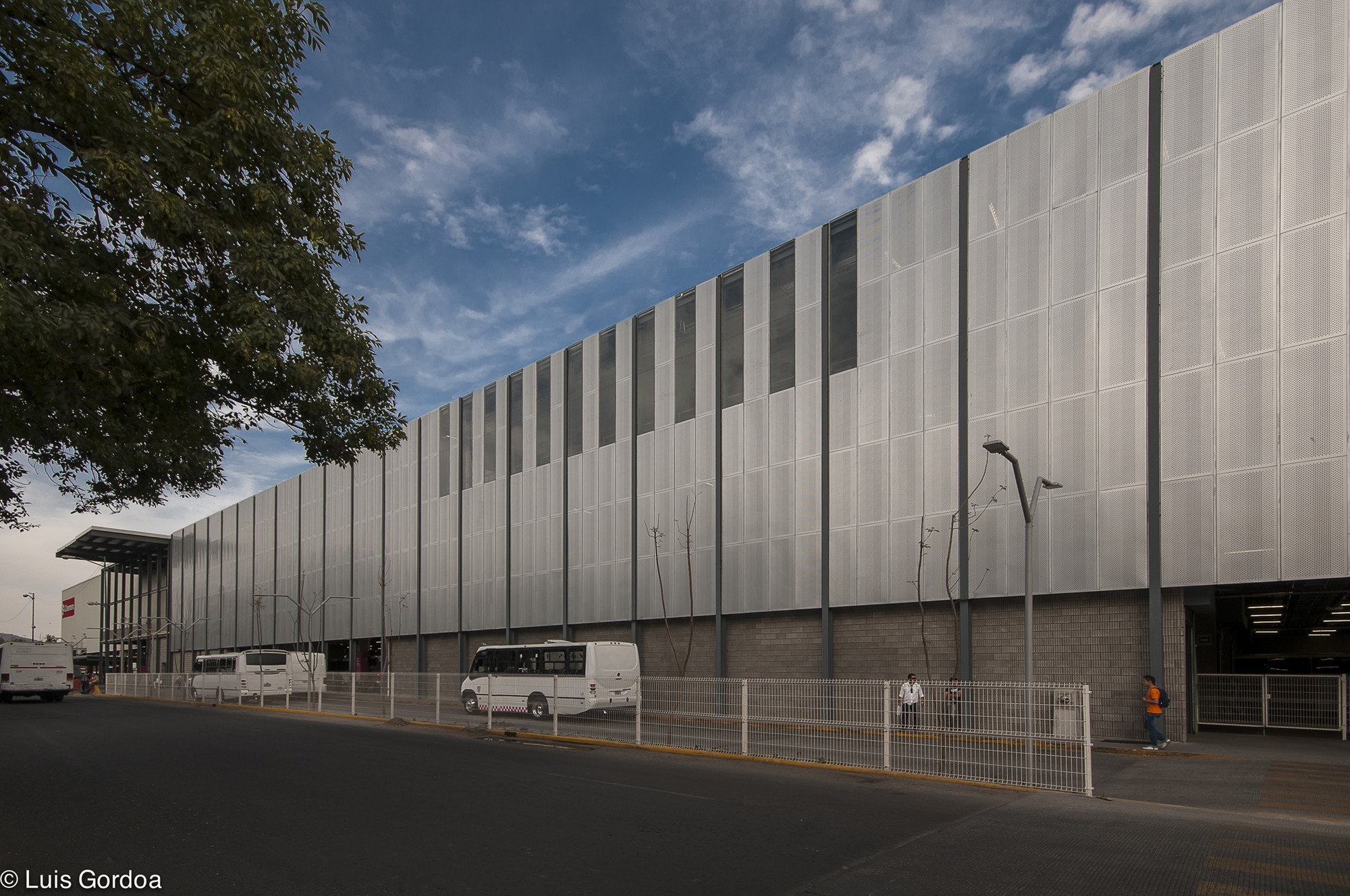 Long perforated metal panel facade above a gray block base, with a white minibus parked at the fence under scattered clouds