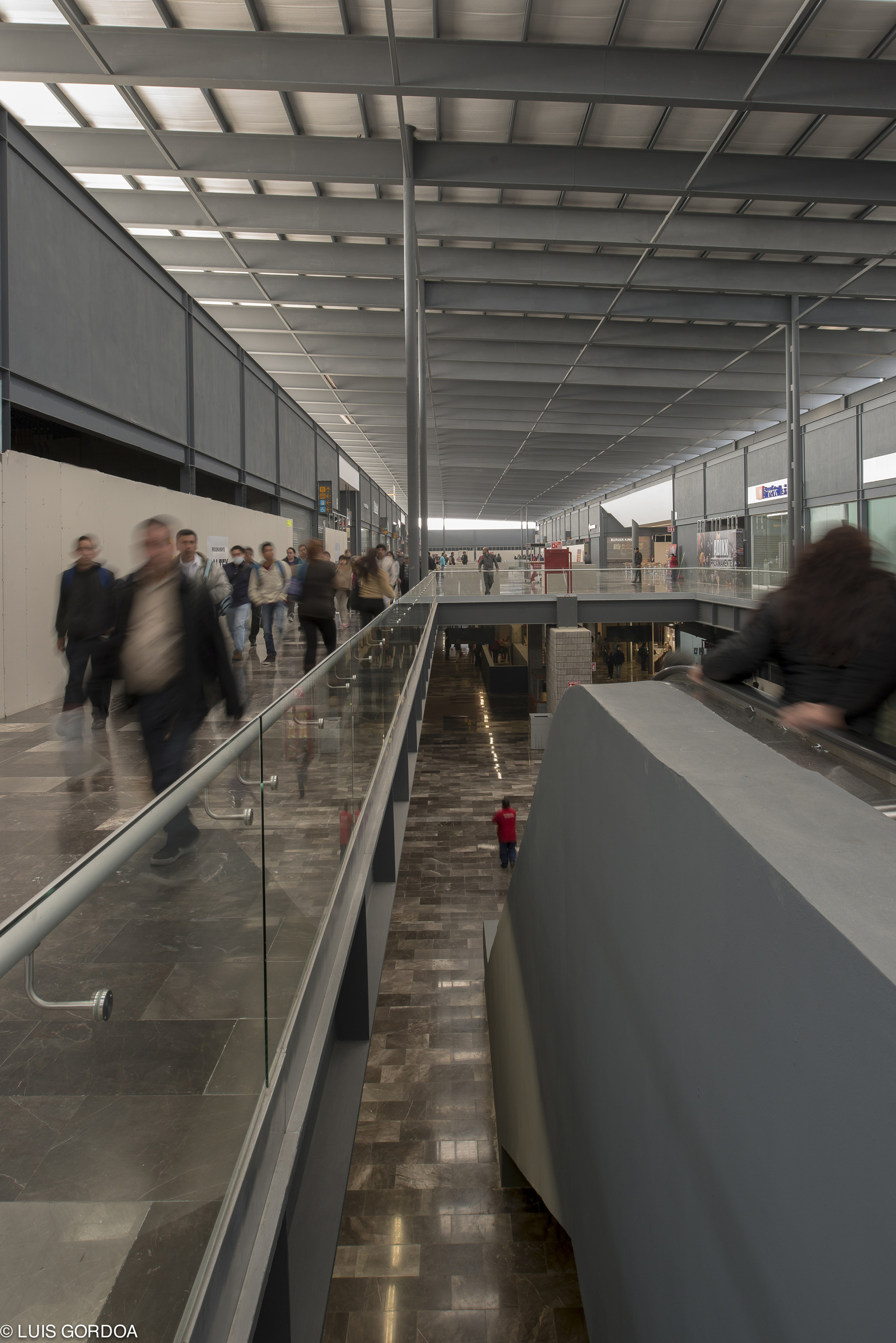 Upper walkway with glass balustrade overlooking a stone-floored concourse, commuters blurred in motion beneath a steel-framed skylit roof