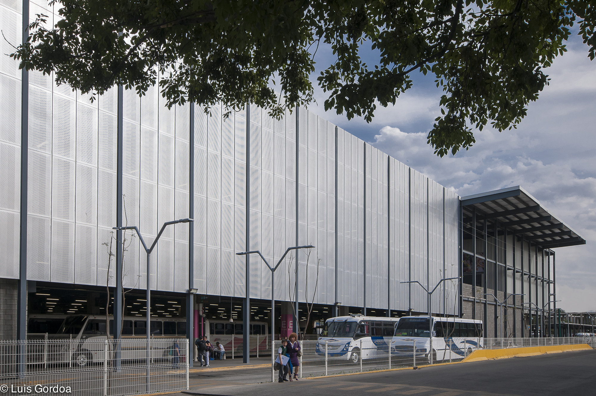 Perforated metal screen facade with slender steel columns and a canopy corner, buses parked below and tree branches overhead