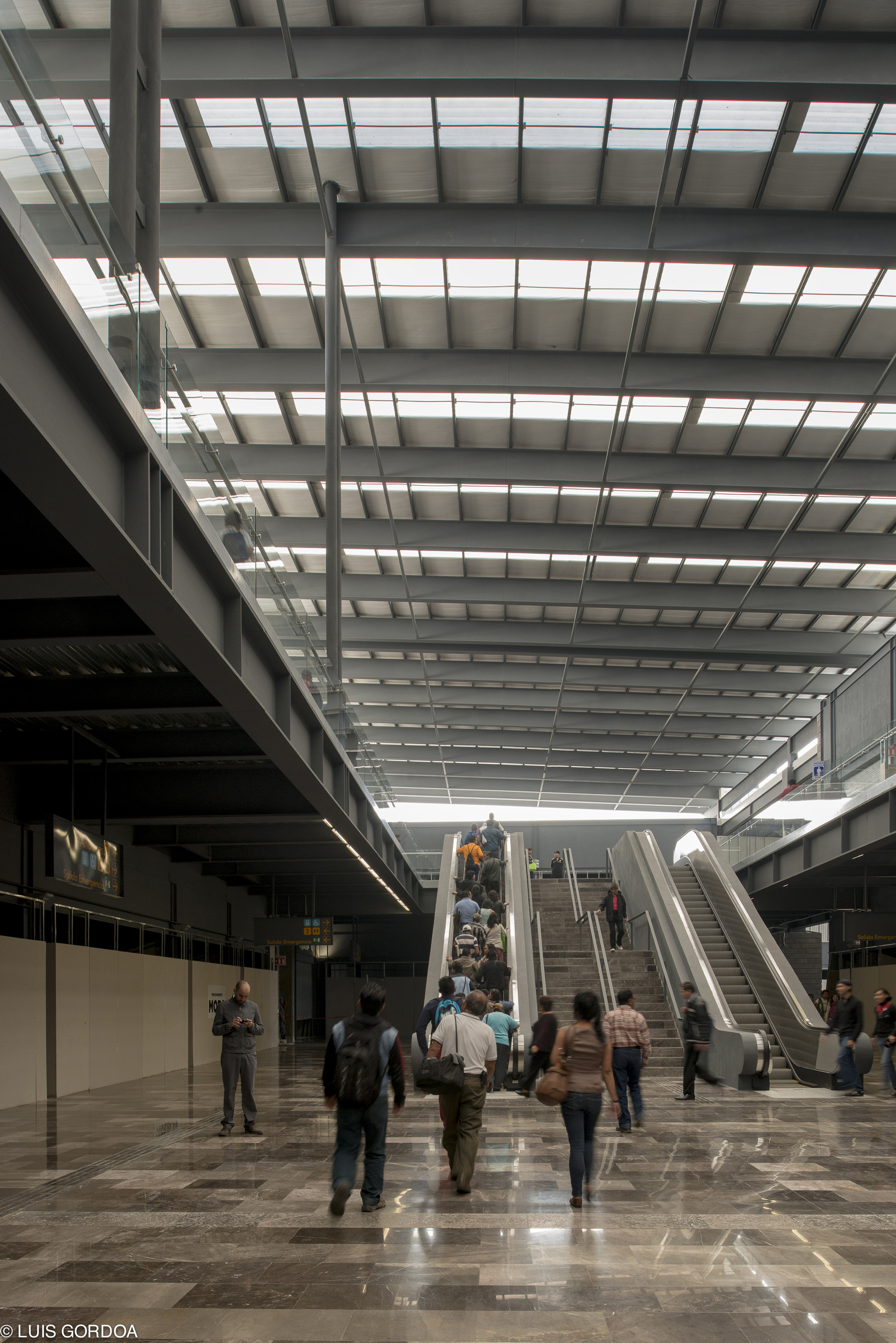 Interior concourse with escalators rising toward a skylit steel roof, travelers crossing the polished stone floor