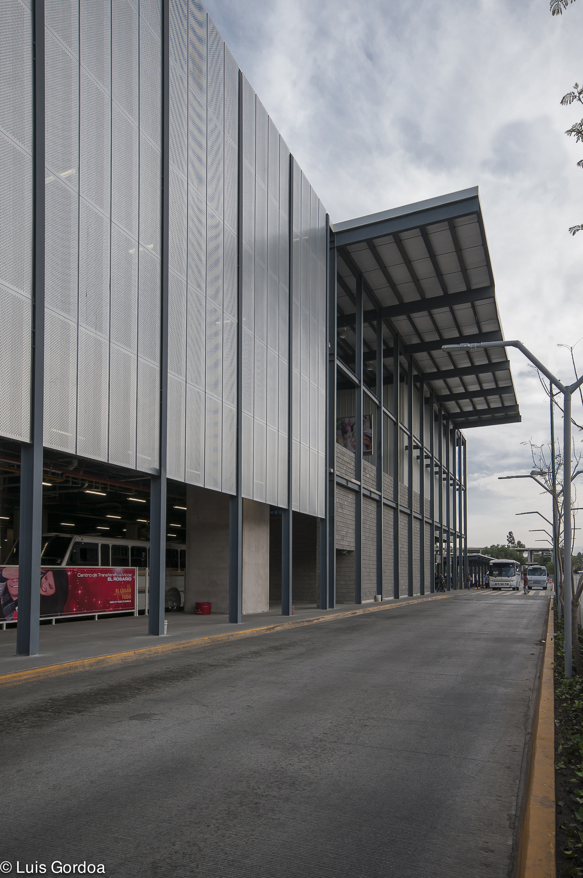 Street-side view of the metal-clad facade and cantilevered steel canopy, a bus stop lane receding under a cloudy sky