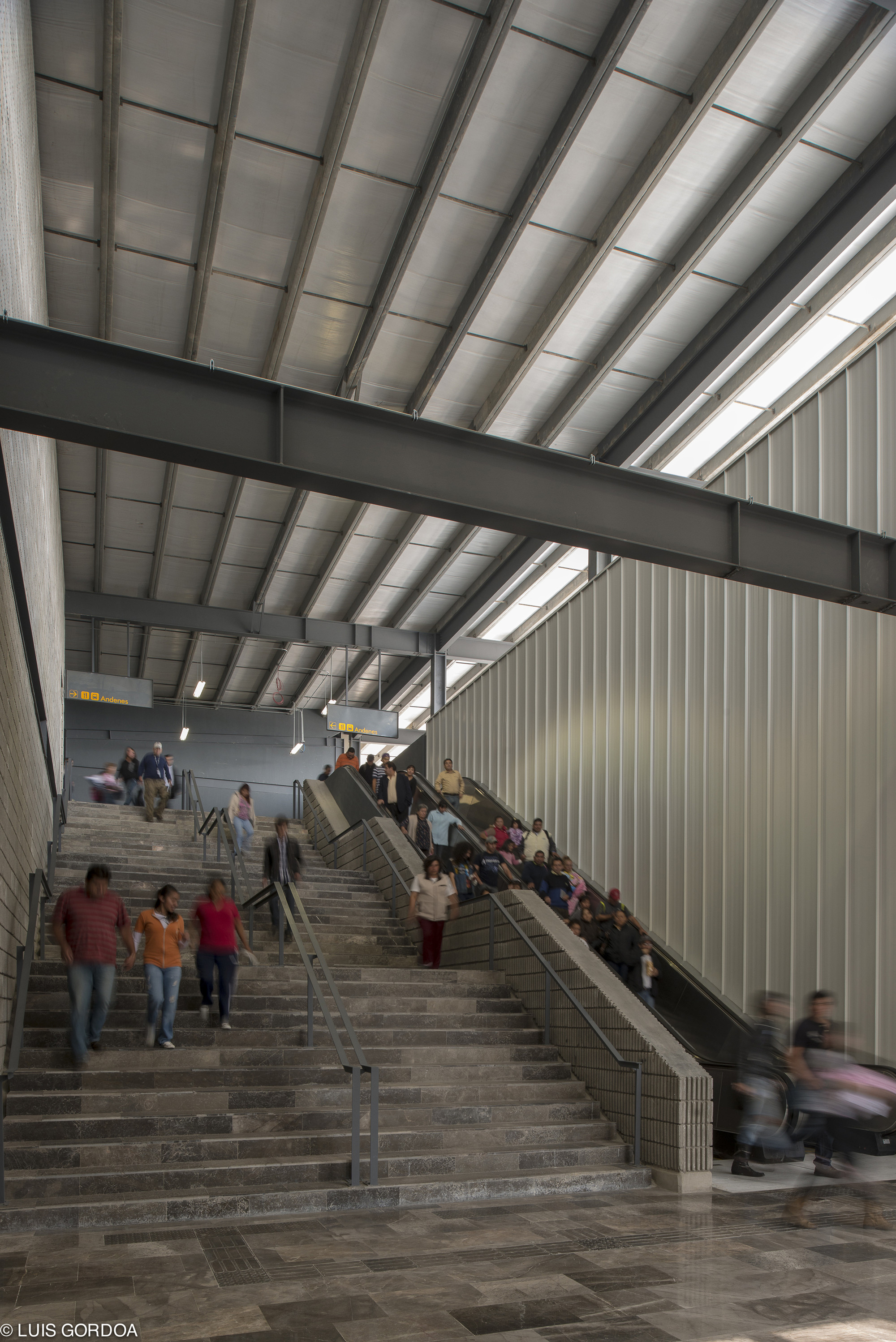 Wide stone staircase and escalators beneath exposed steel beams and a sloped skylit ceiling, commuters climbing and descending