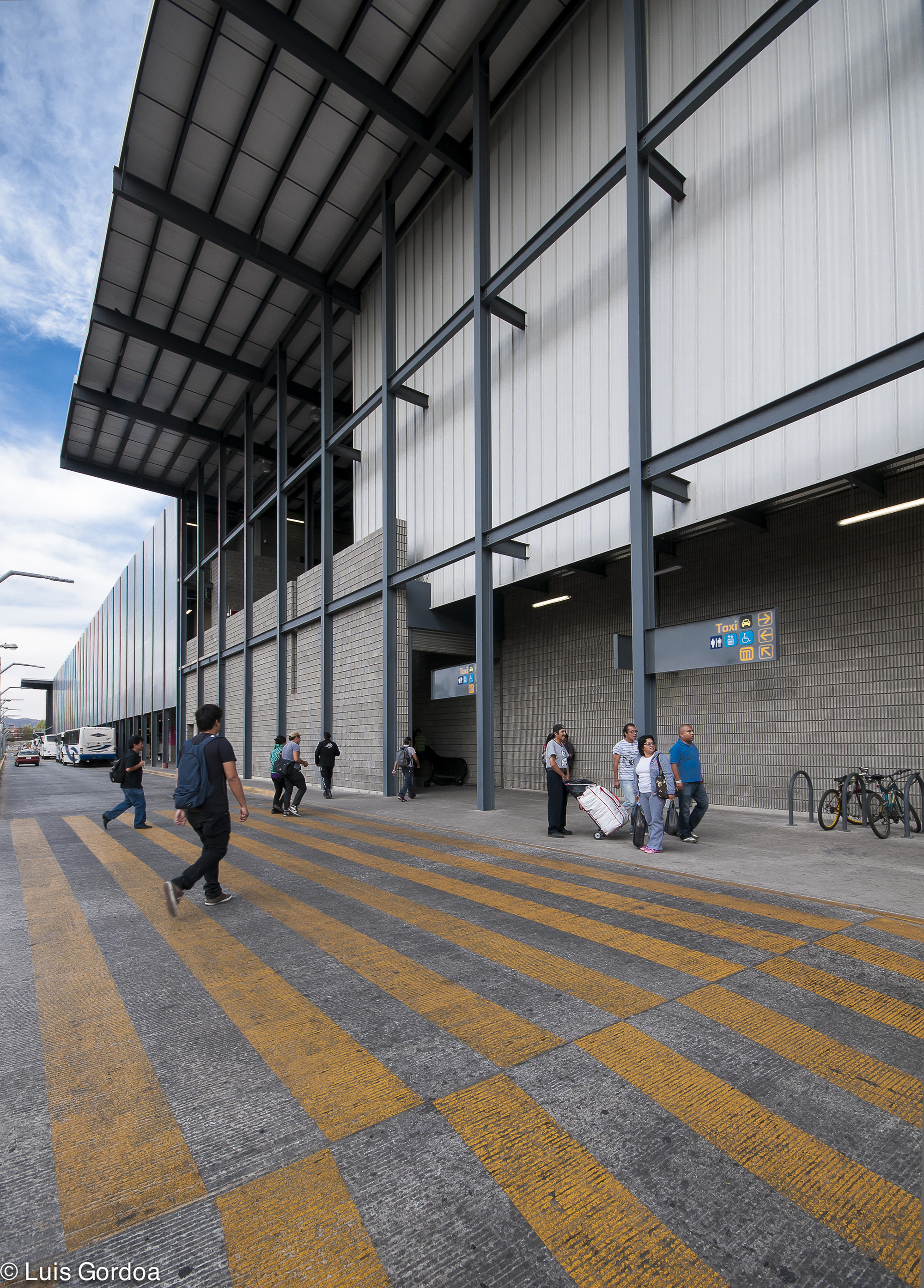 Steel-framed station facade with corrugated metal cladding and block base, seen across a yellow-striped crosswalk with travelers and parked bicycles under a par