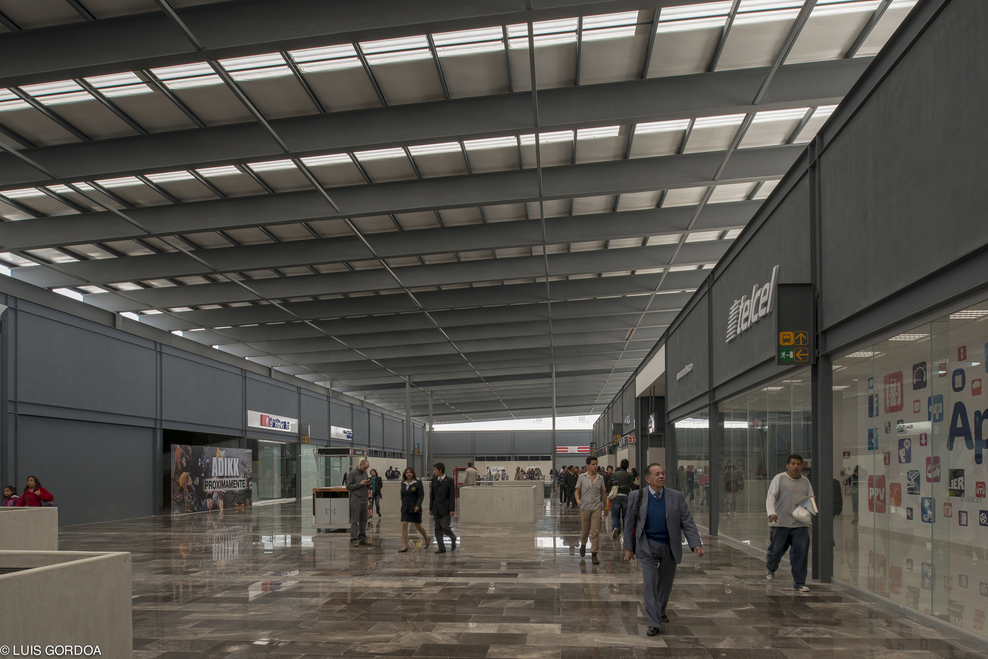Long concourse with a skylit steel roof grid, polished stone floor and shopfronts, with commuters walking toward the far end