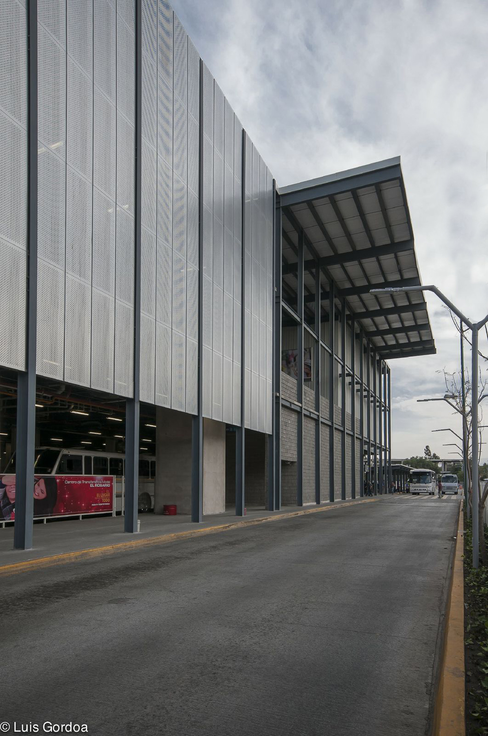 Perforated metal screen facade beside a cantilevered steel canopy, with buses parked below and a road under an overcast sky