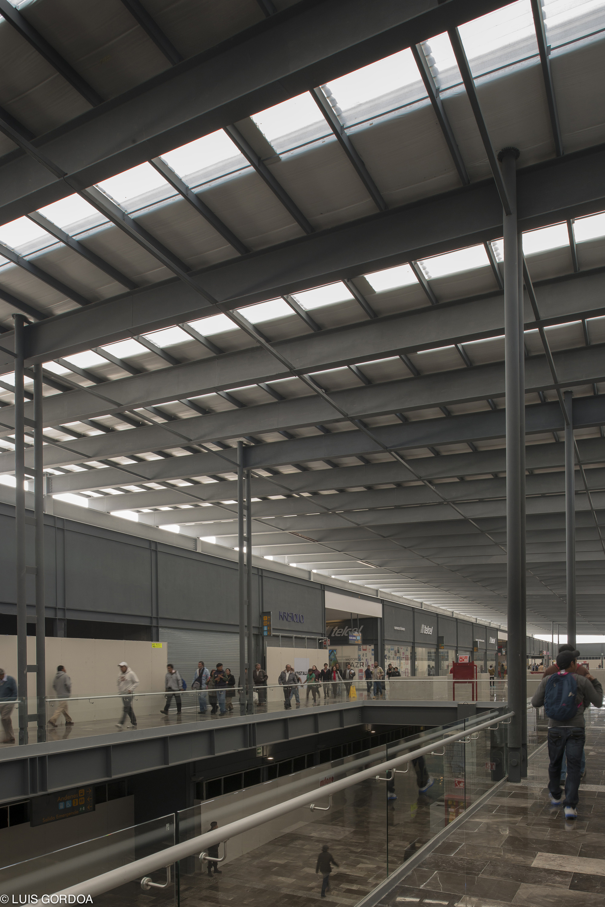 Upward view of skylit steel roof beams and slender columns above a mezzanine walkway where commuters pass behind glass railings